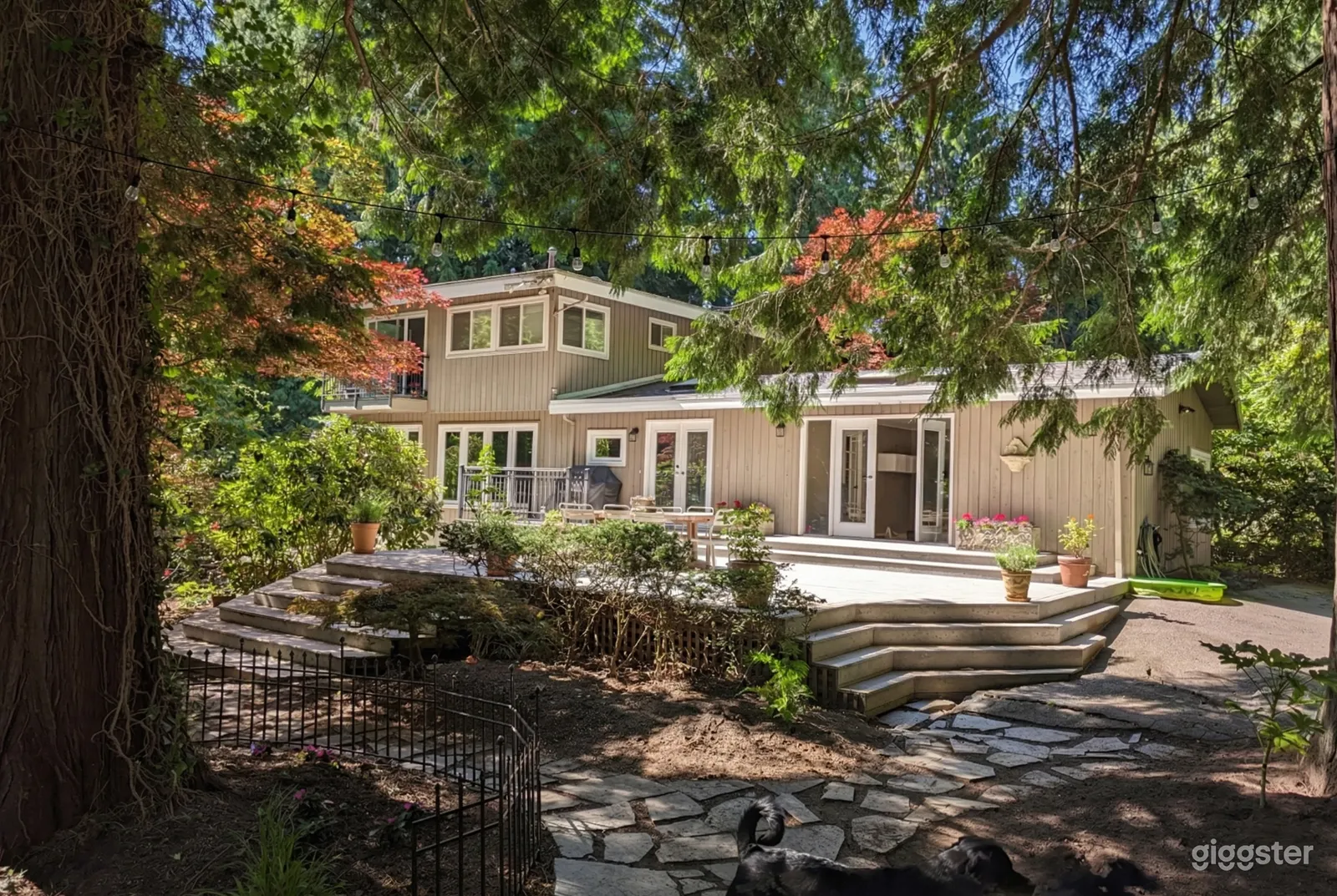A long modern home with many windows overlooks a large deck and wooded garden area.
