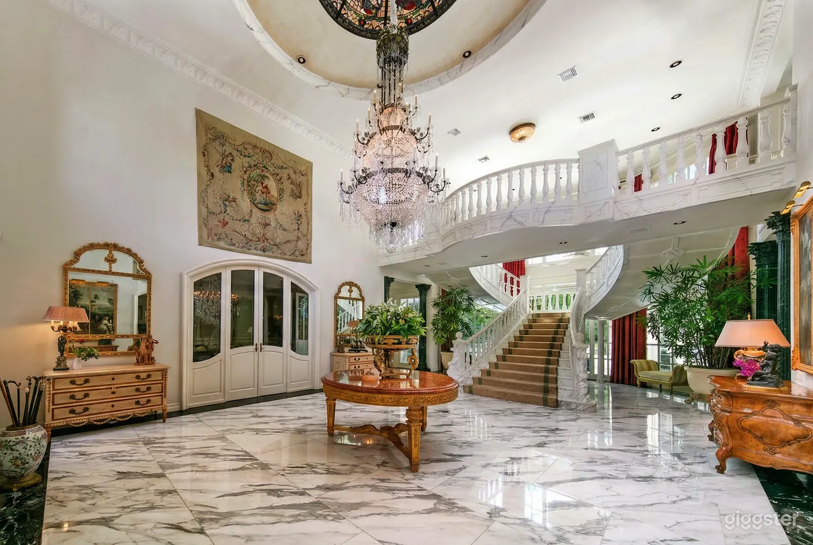 A wide staircase sits under a balconied second floor in a ballroom venue with ceramic floor and ceiling chandelier.