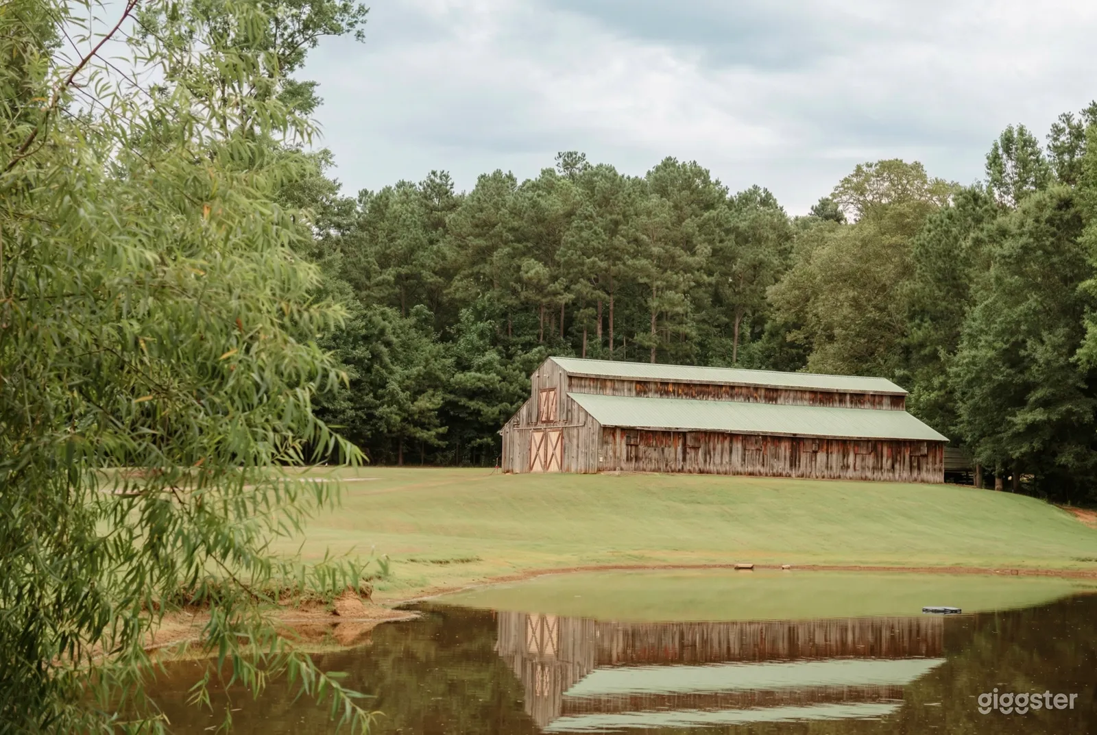 A long two floored barn with closed doors sits in a freshly mowed field surrounded by tall trees.