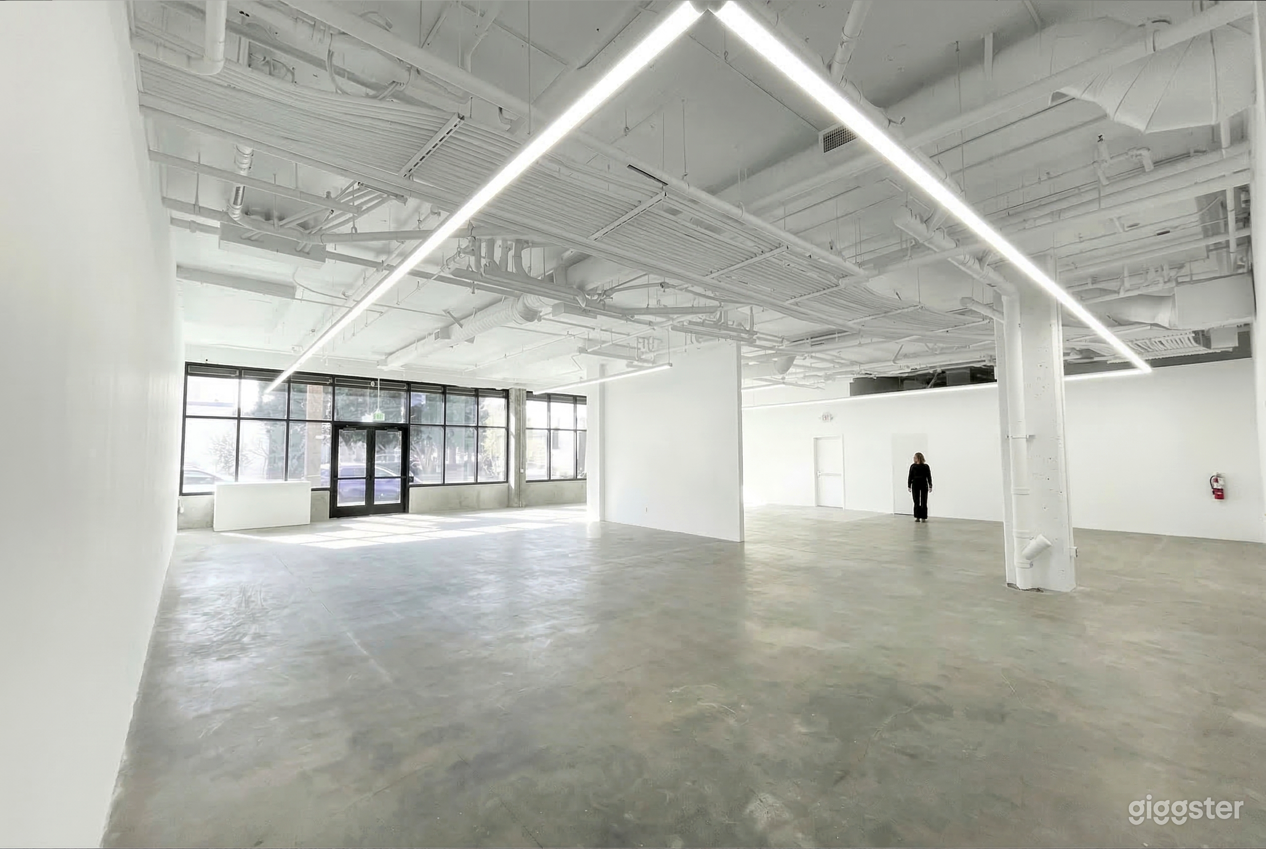 An empty white warehouse type space with one wall of windows and an industrial type ceiling painted white.