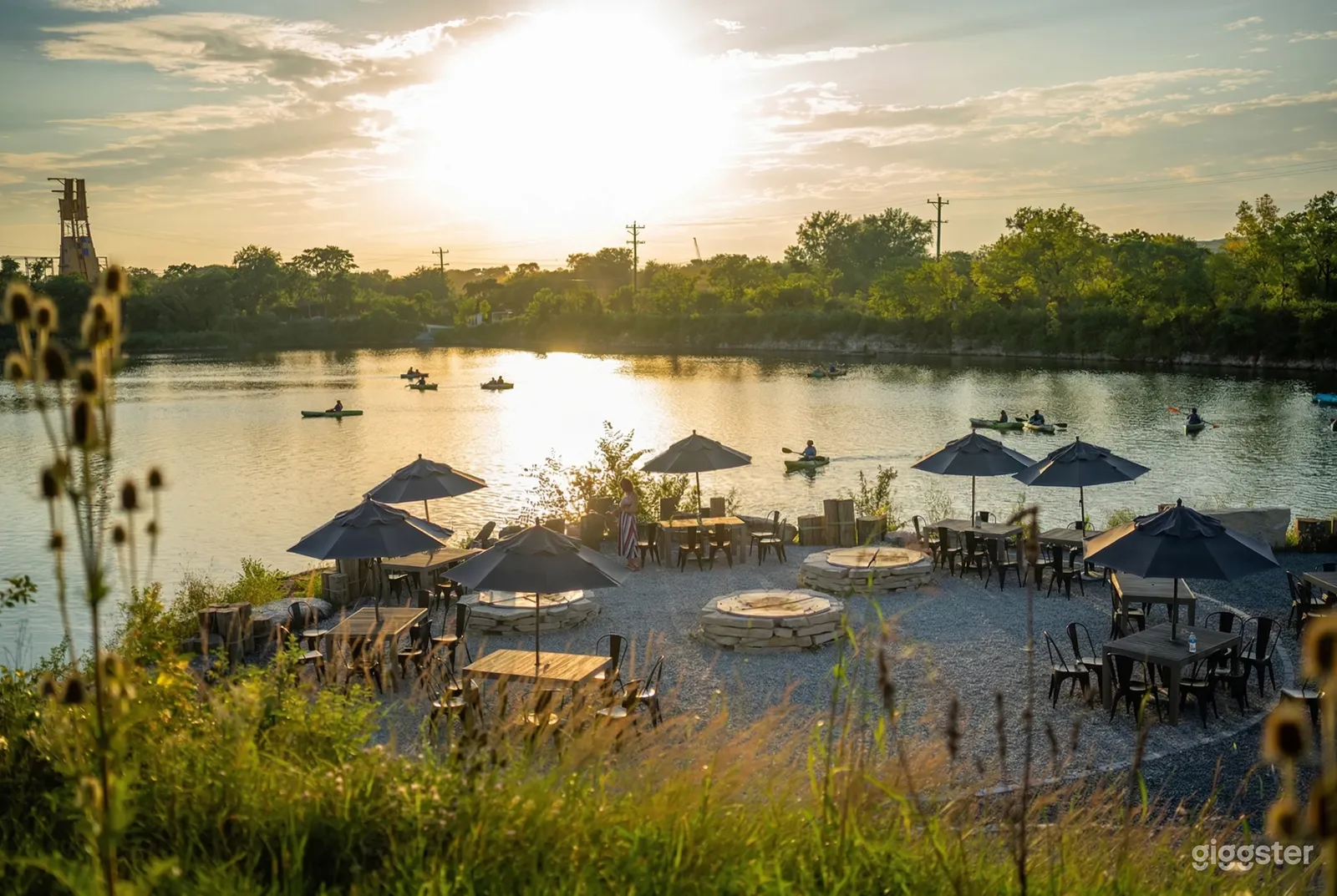 Umbrella covered tables and stone firepits sit beside a lake where people are canoeing.