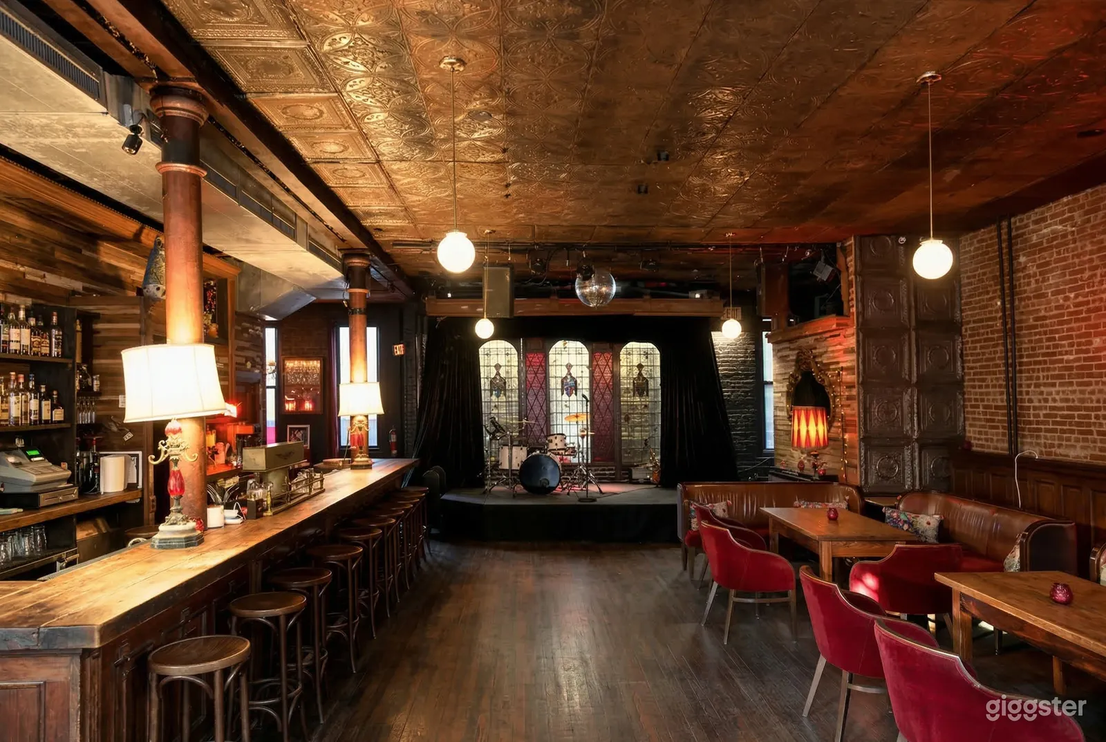 Stools line a well lit bar with a drum kit on a stage in a traditionally styled eatery.