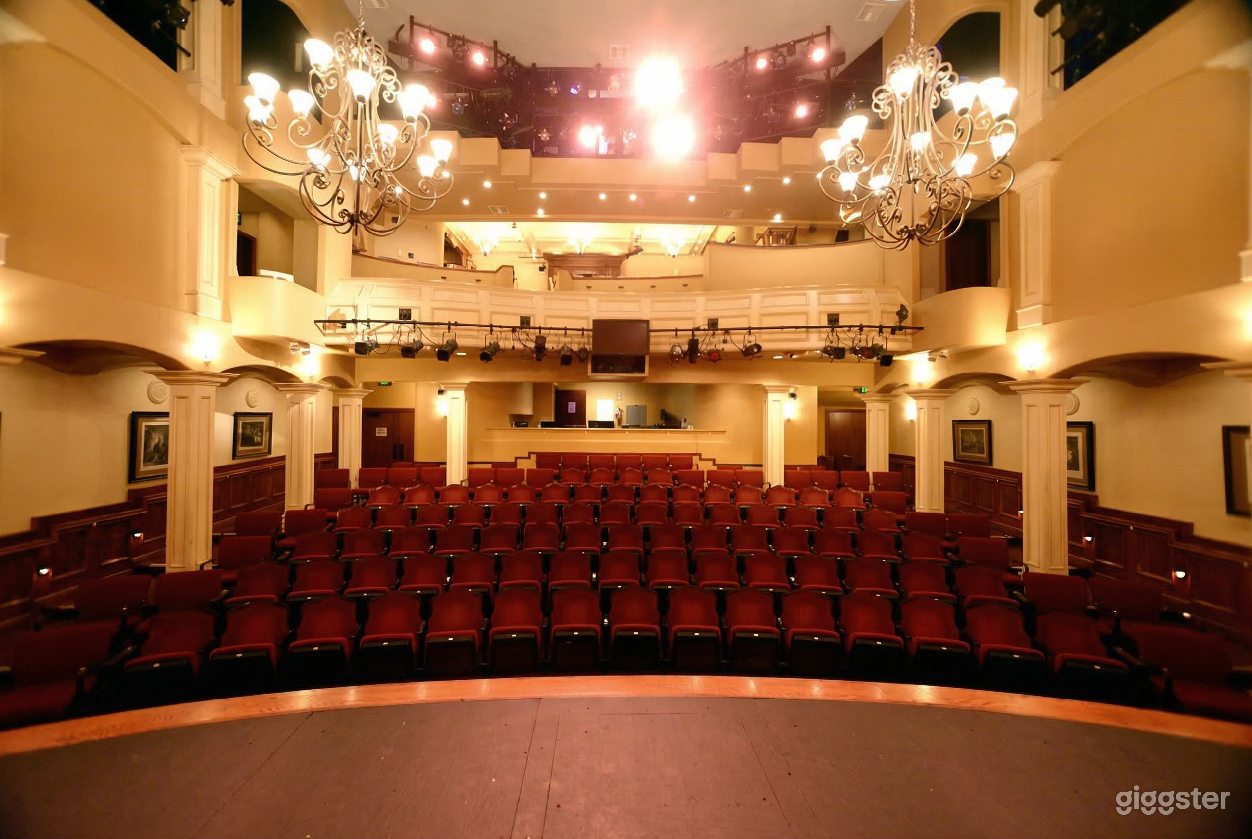 Ornate chandeliers hang over cushioned theater stairs in front of a stage.