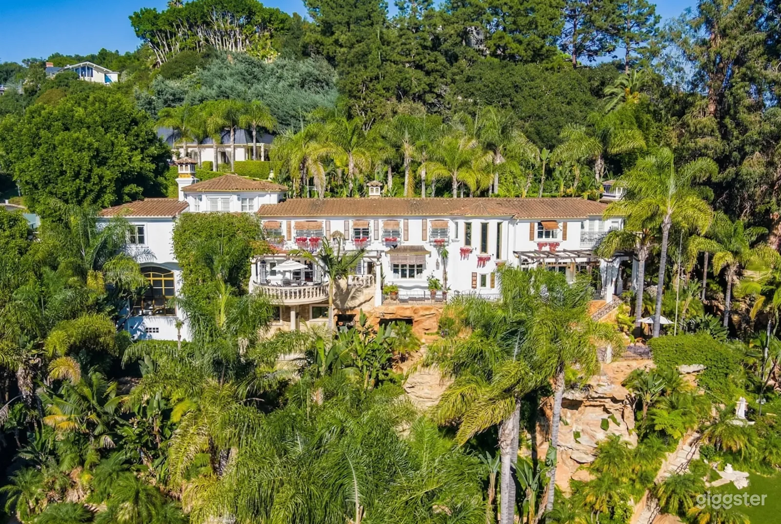 An expansive Tuscan style villa sits on a hillside and is surrounded by palms and other types of trees.