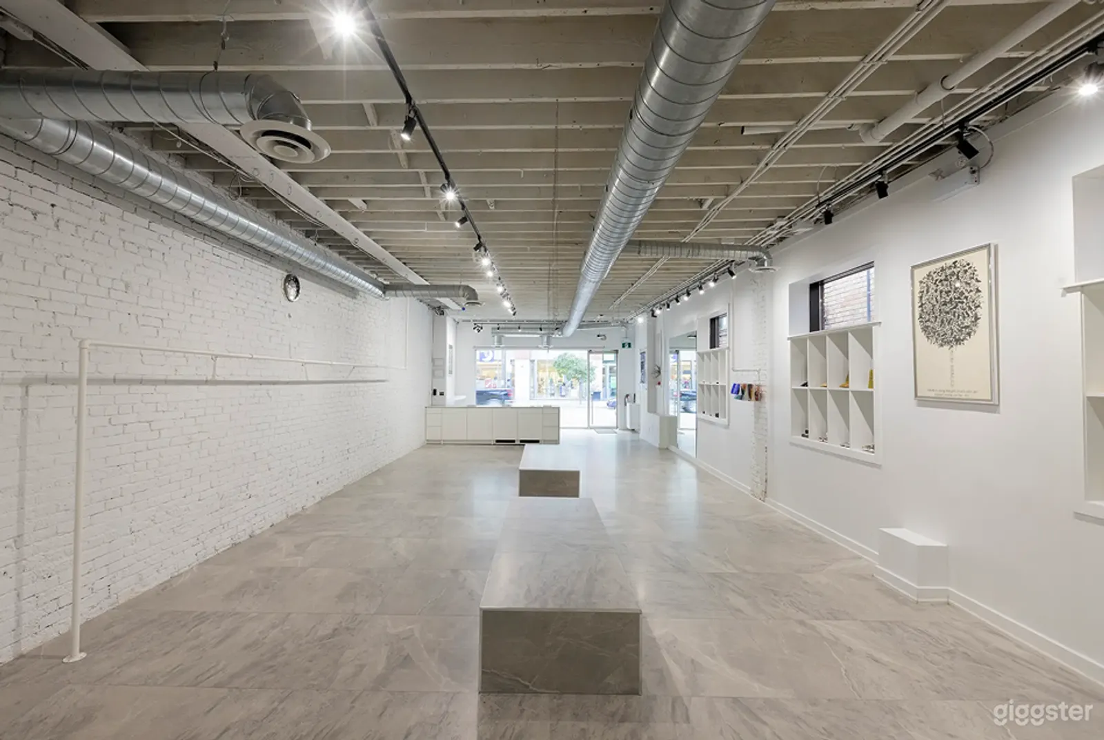 A room with silver industrial piping along the ceiling, white brick and wood walls, and floor to ceiling window and door at the end. 
