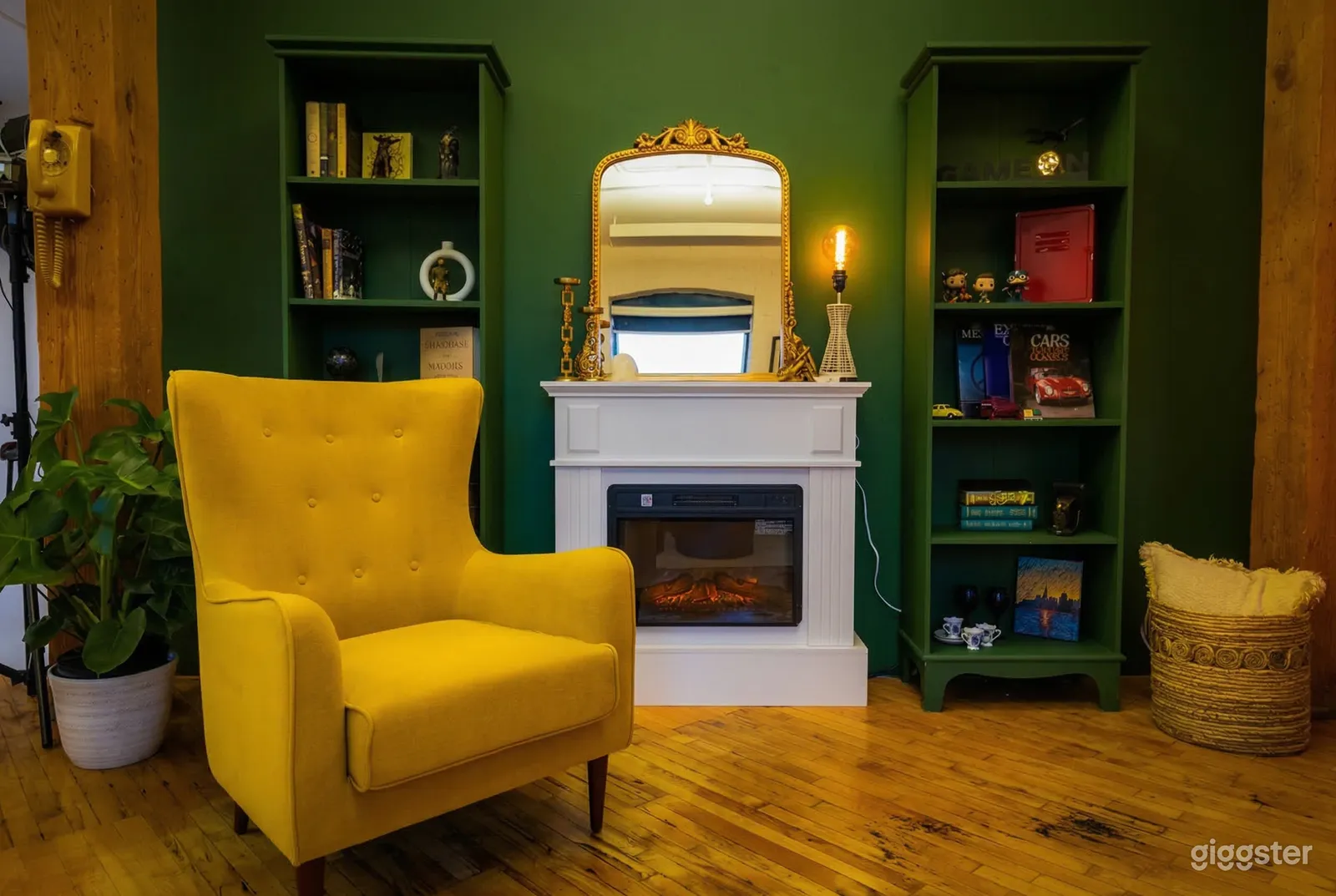 A bright yellow wing back chair sits in front of an emerald green wall with shelving and a white fireplace.