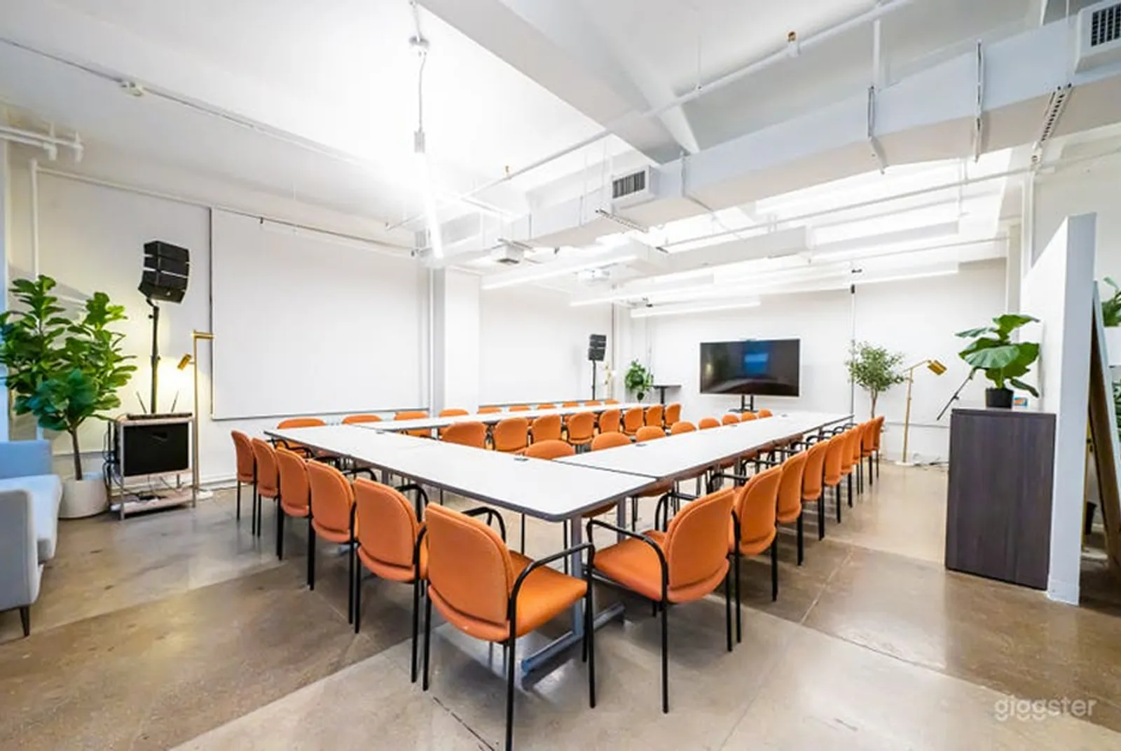 A sizeable U shaped table with many orange chairs sits in front of a large black tech screen.