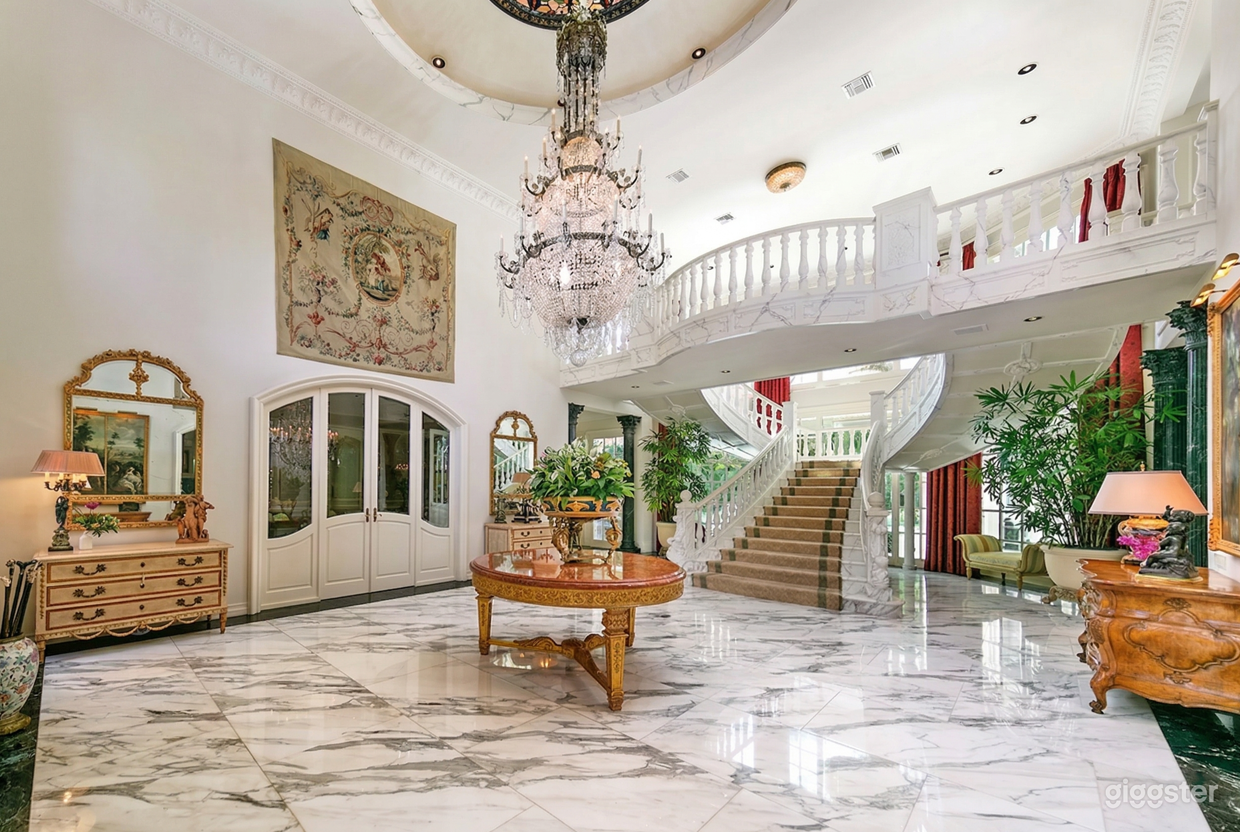 A bright ballroom style room has a wide central staircase and a balcony overlooking a piazza with chandelier.