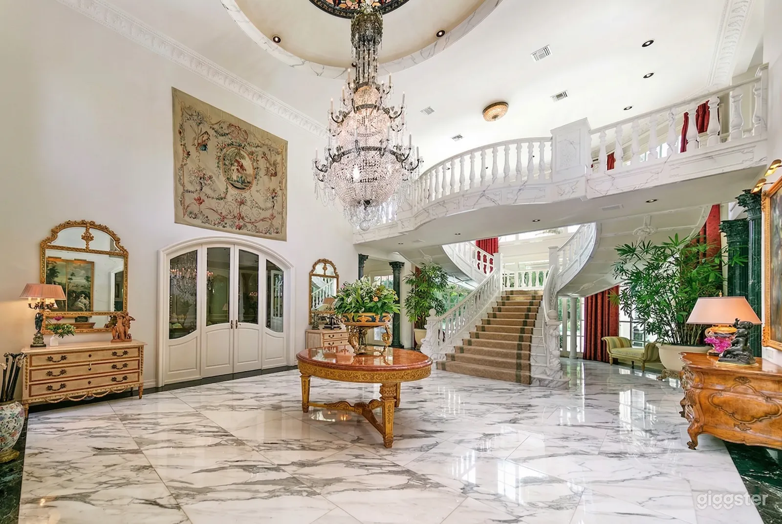 A bright ballroom style room has a wide central staircase and a balcony overlooking a piazza with chandelier.