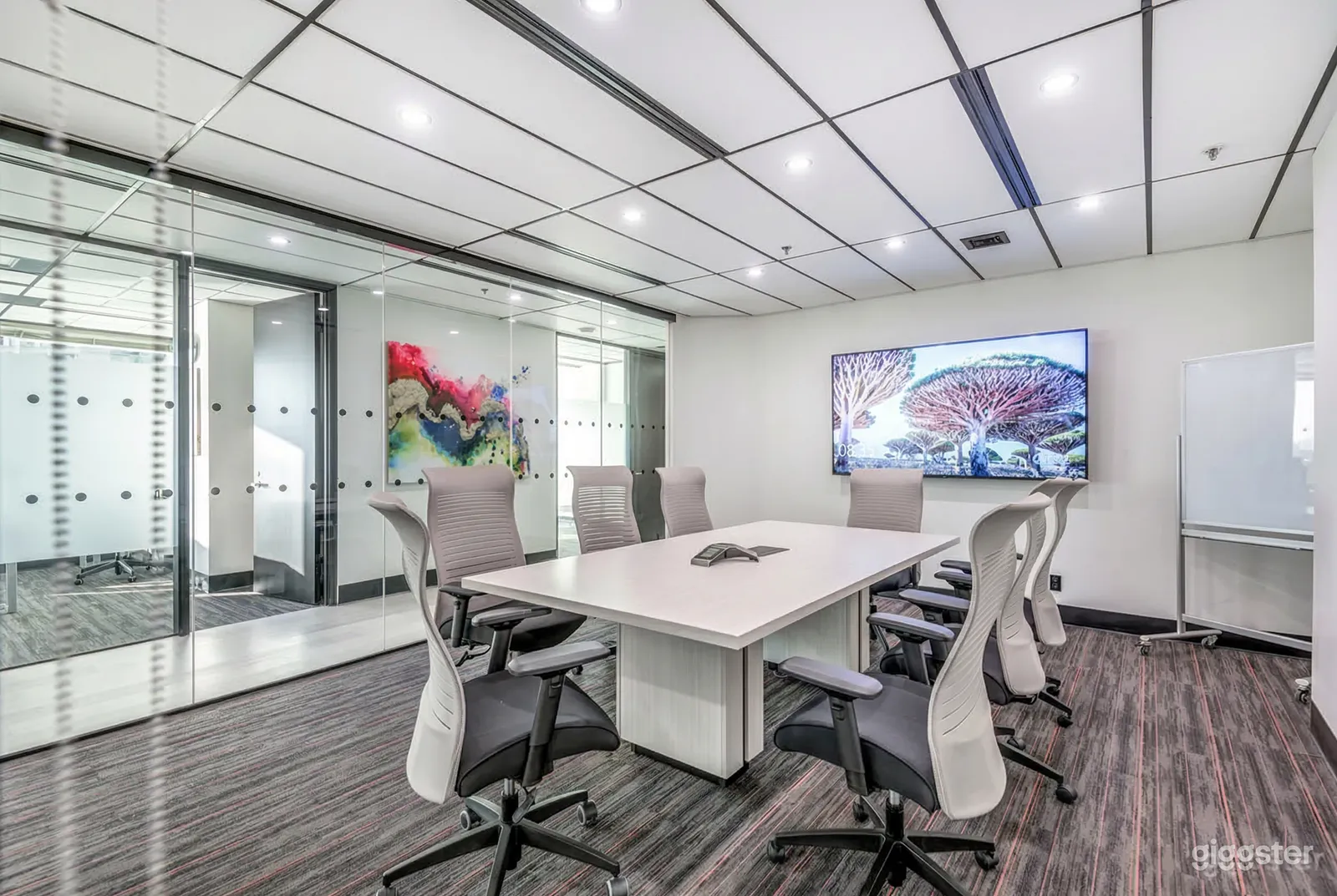 A conference room with a glass wall contains a white conference table and ergonomic chairs in front of a tech screen.