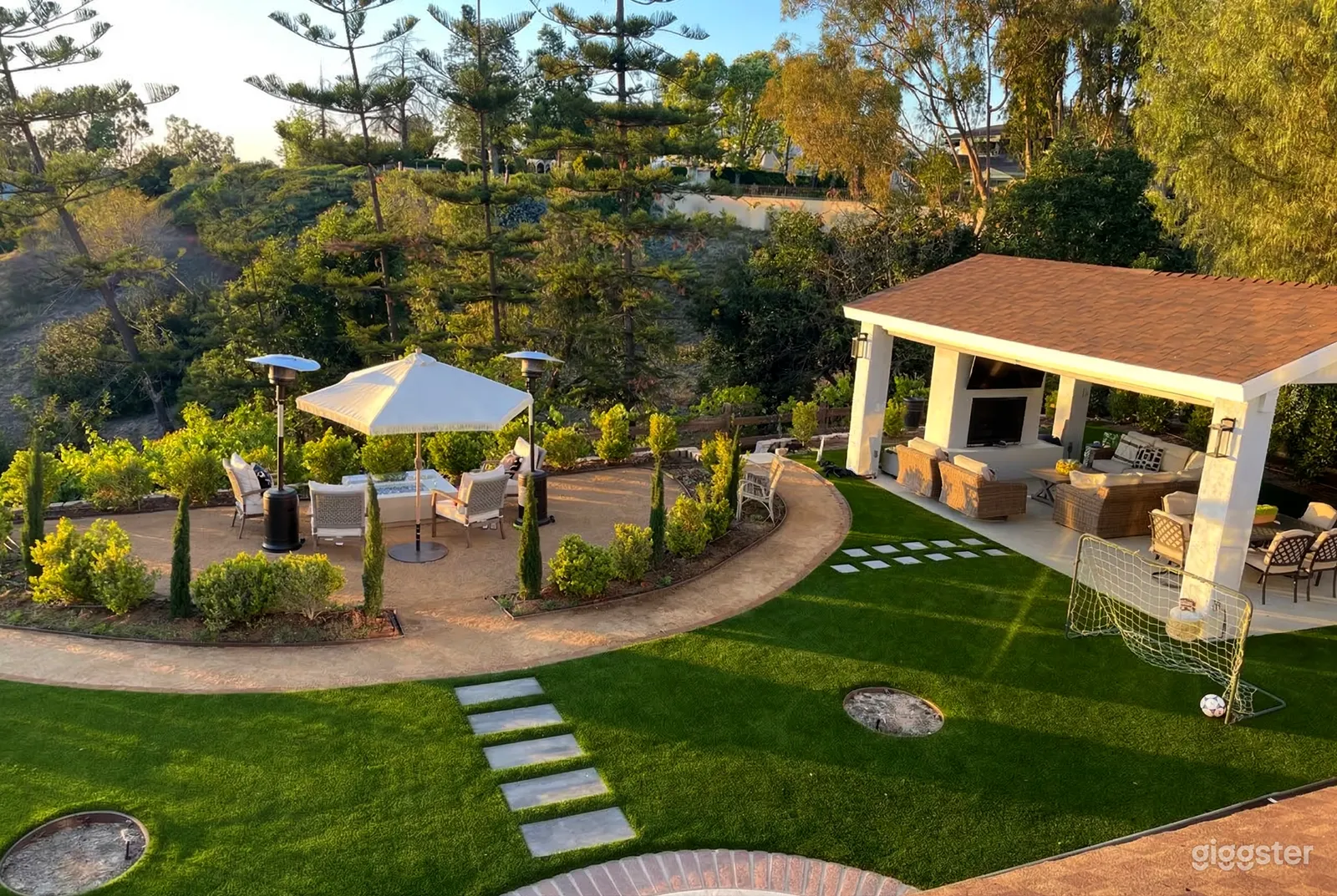 A stone patio with seating and hedges sits to the left of a painted cement cabana with TV and fireplace.