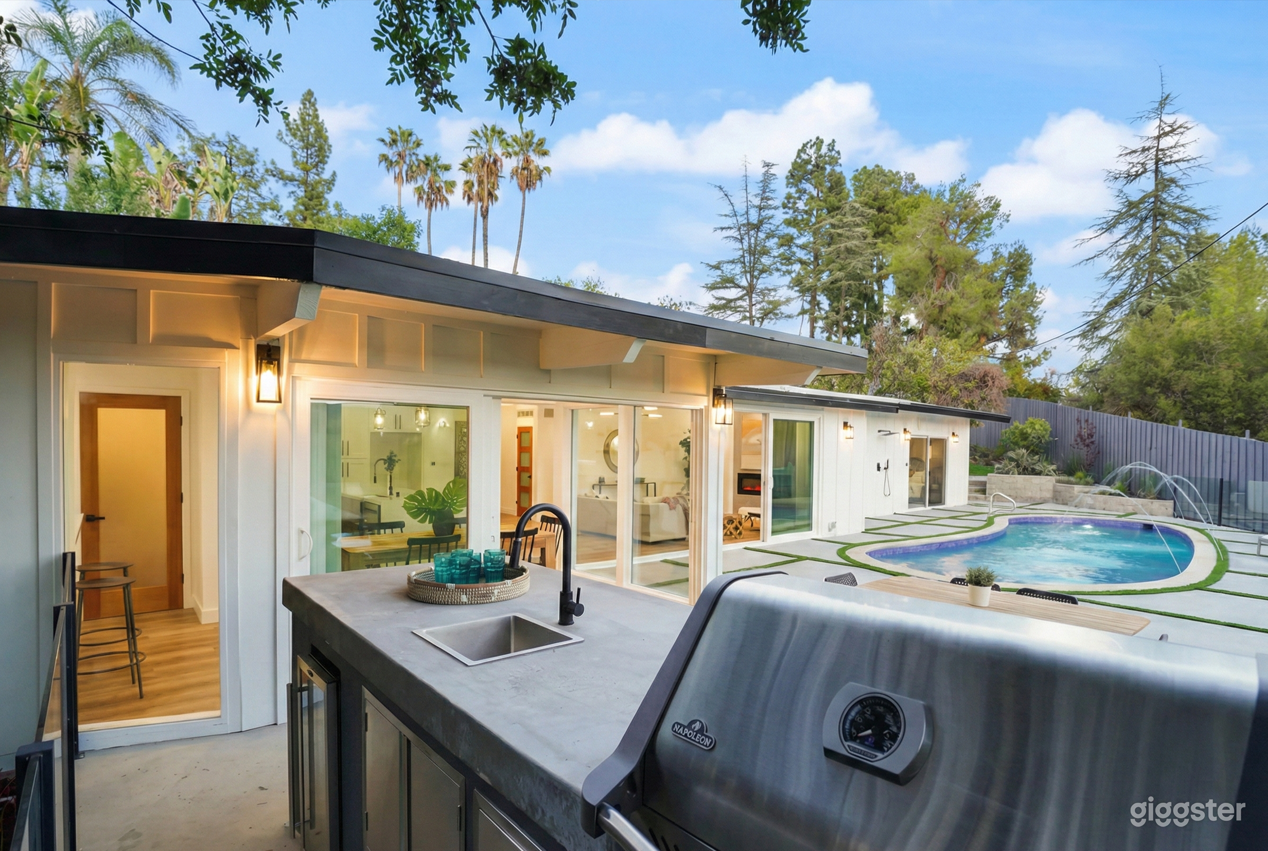 White exterior and window filled open concept bungalow facing a pool and outdoor kitchen.
