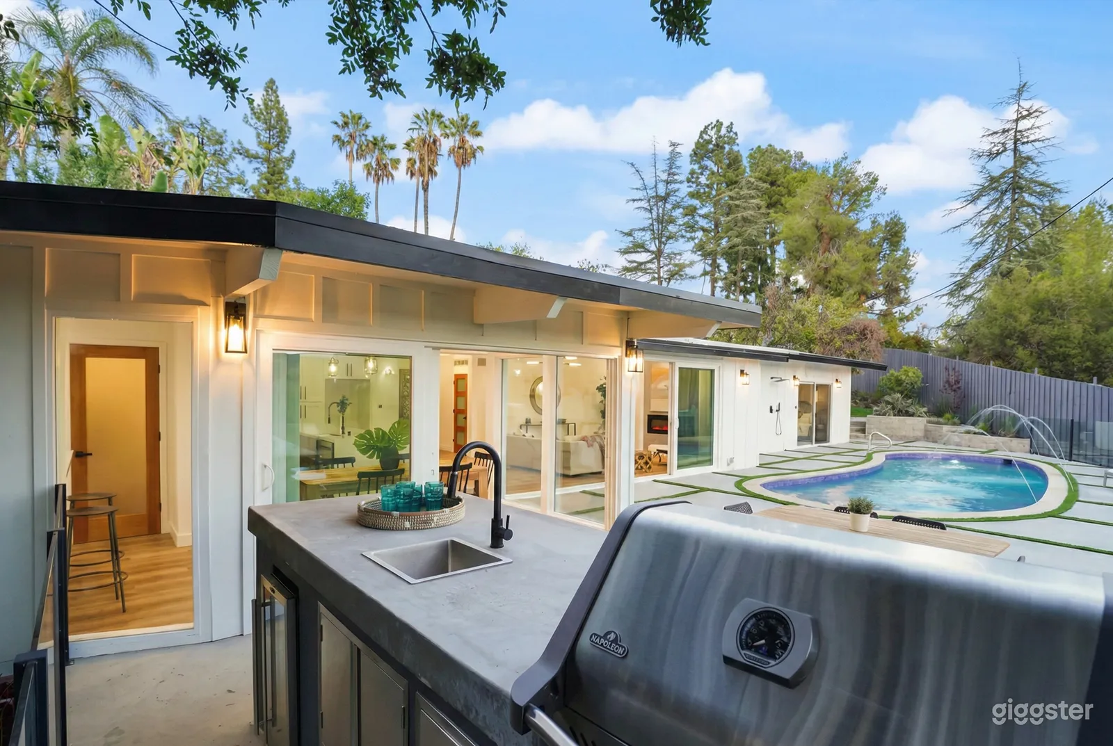 White exterior and window filled open concept bungalow facing a pool and outdoor kitchen.