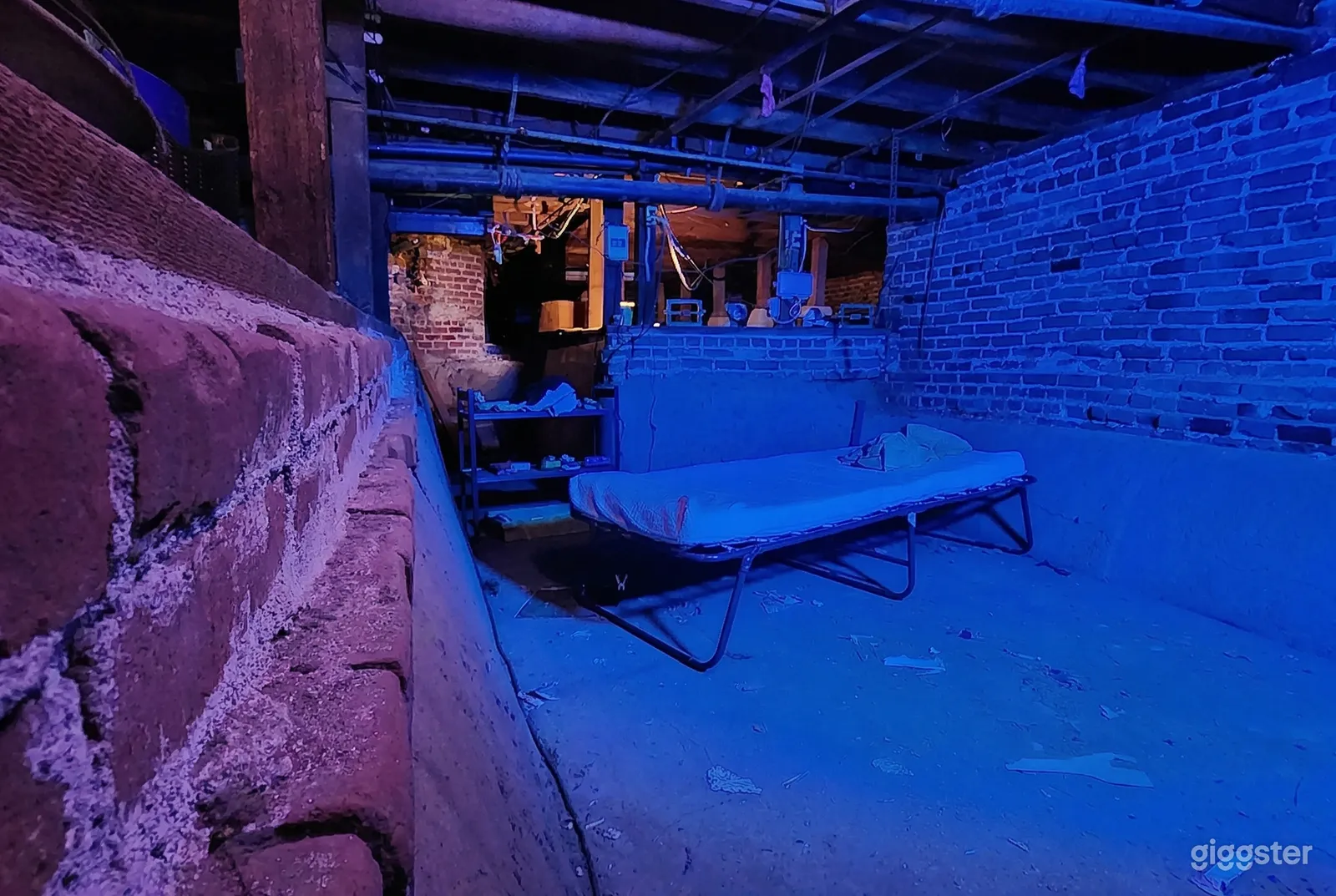 A basement setting with brick walls, cement floor, and cot in the middle of the room is lit in blue.