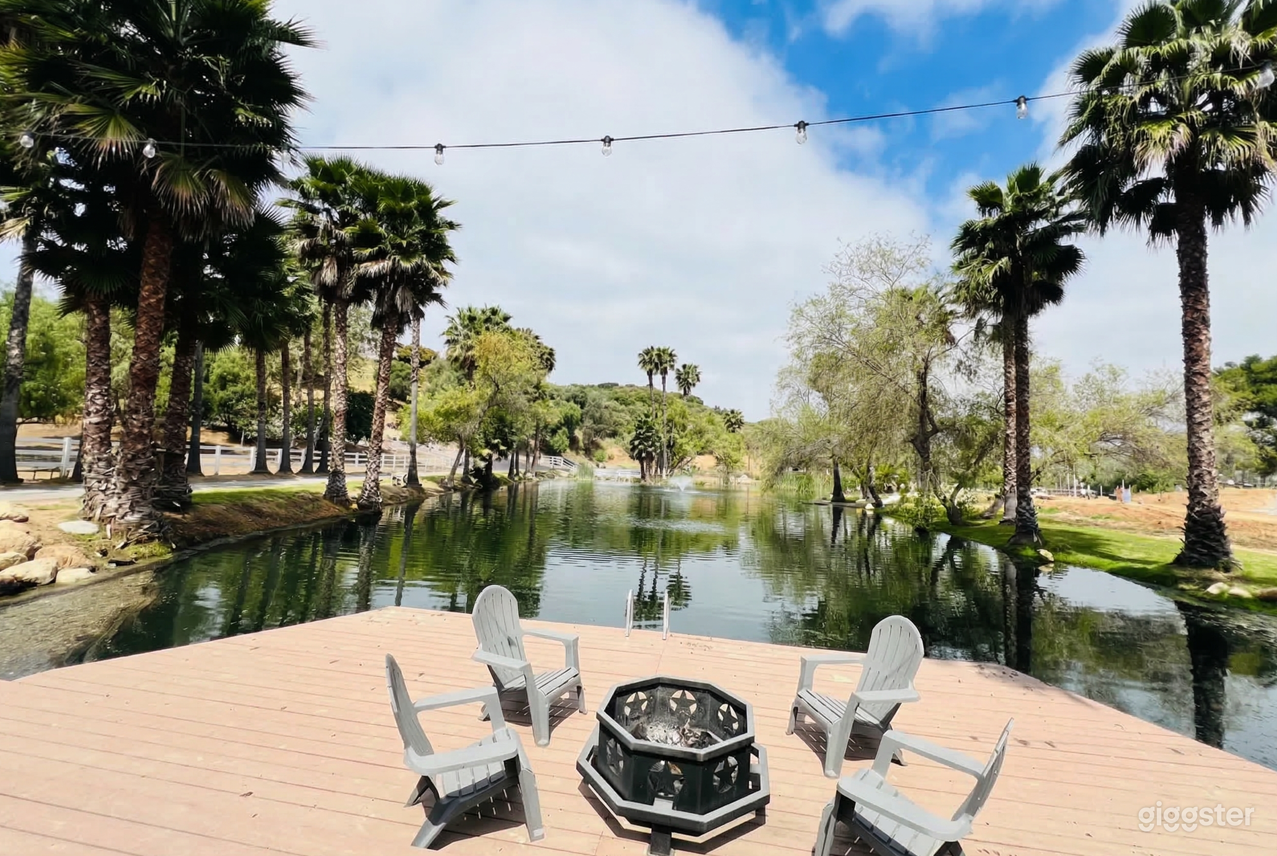 Four chairs surround a fireplace on a deck overlooking a body of water flanked by trees.