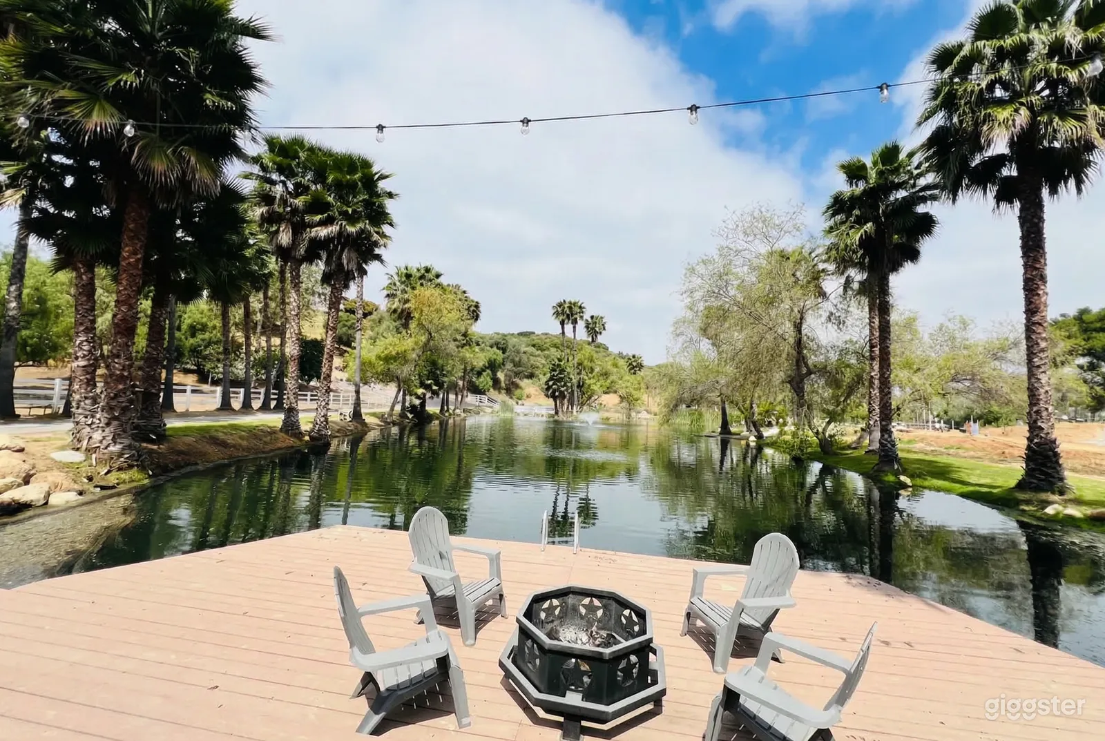 Four chairs surround a fireplace on a deck overlooking a body of water flanked by trees.