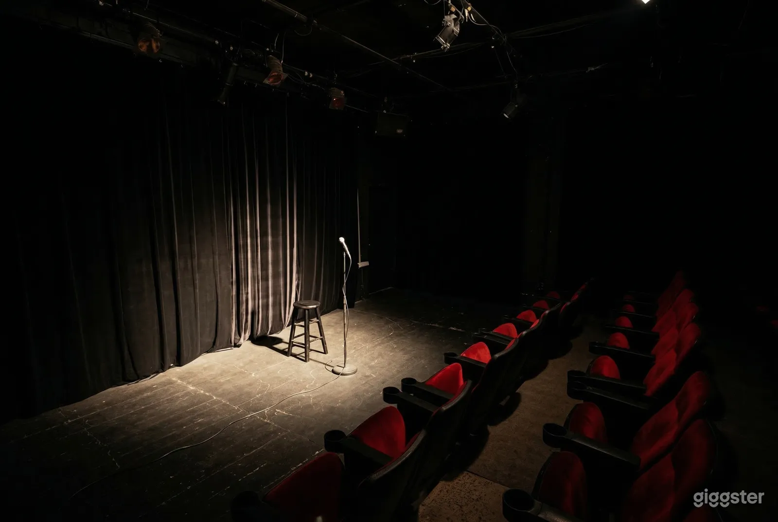 A lone microphone on a stand beside a stool sits in front of curtains on one side and theater seating on the other side.