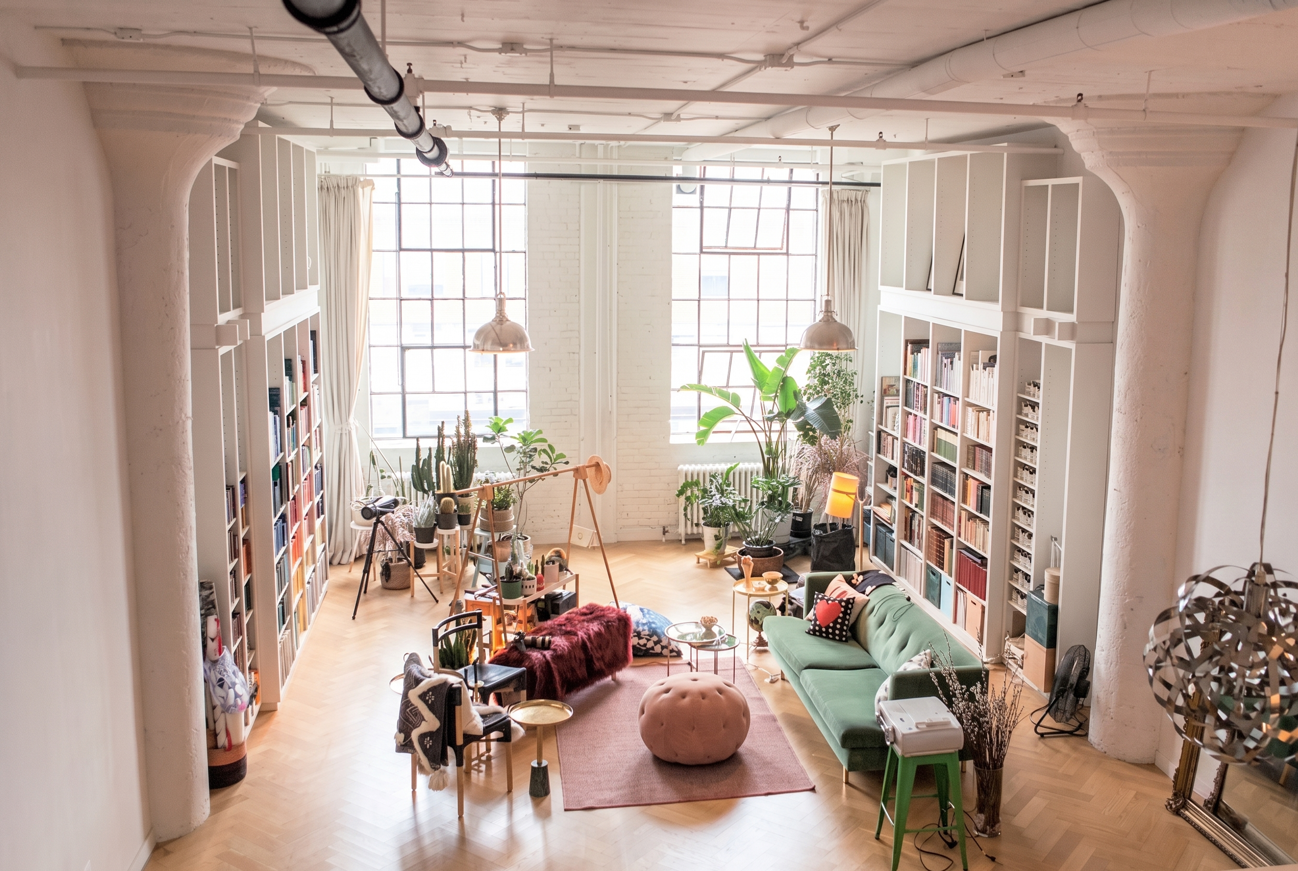 An industrial chic room with a high ceiling has book shelves on both sides, artsy seating in the middle, and large plants in front of the windows.