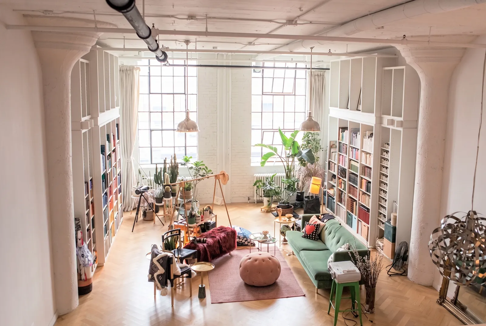 An industrial chic room with a high ceiling has book shelves on both sides, artsy seating in the middle, and large plants in front of the windows.