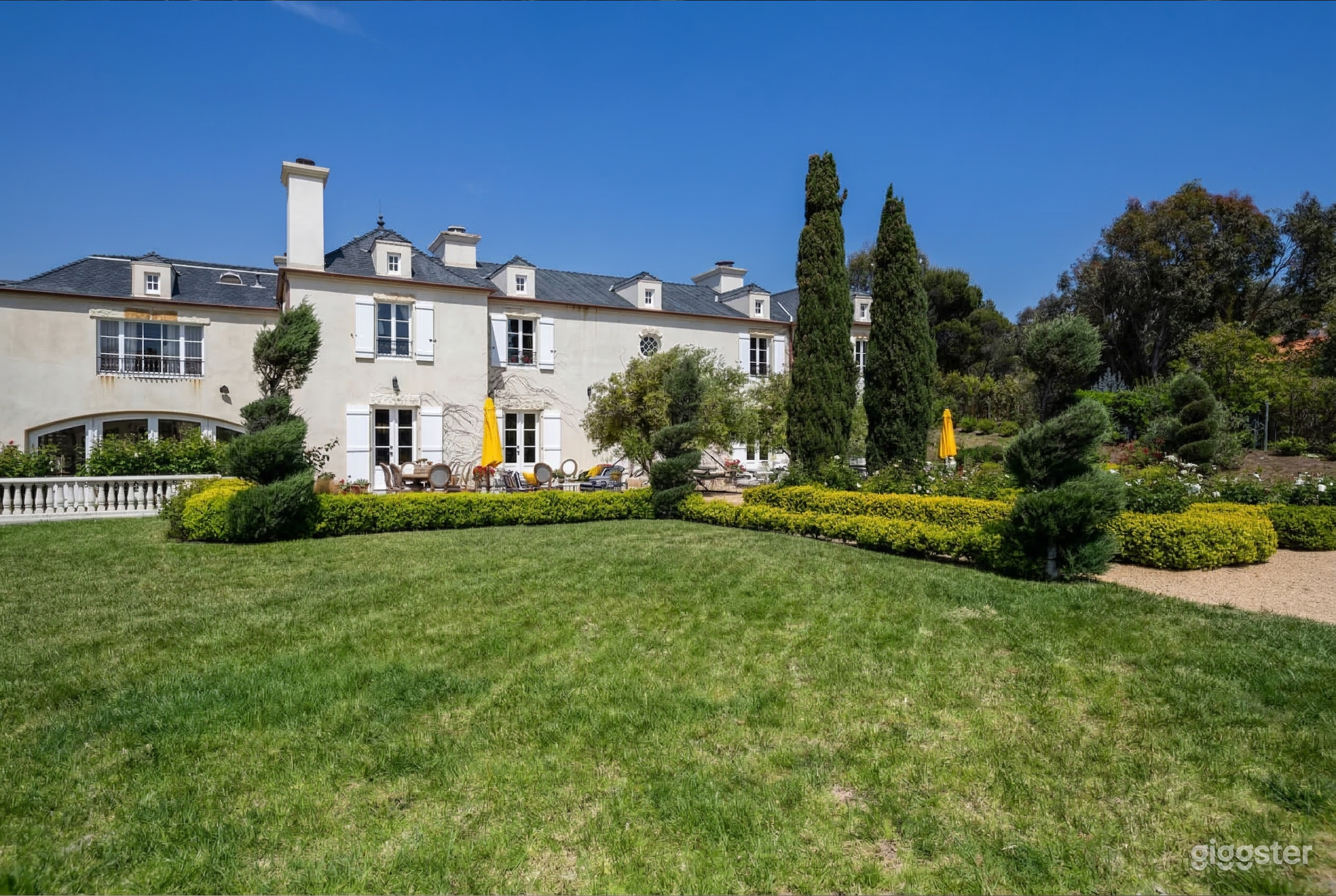 An expansive grey mansion overlooks a large garden with carved hedges and trees.