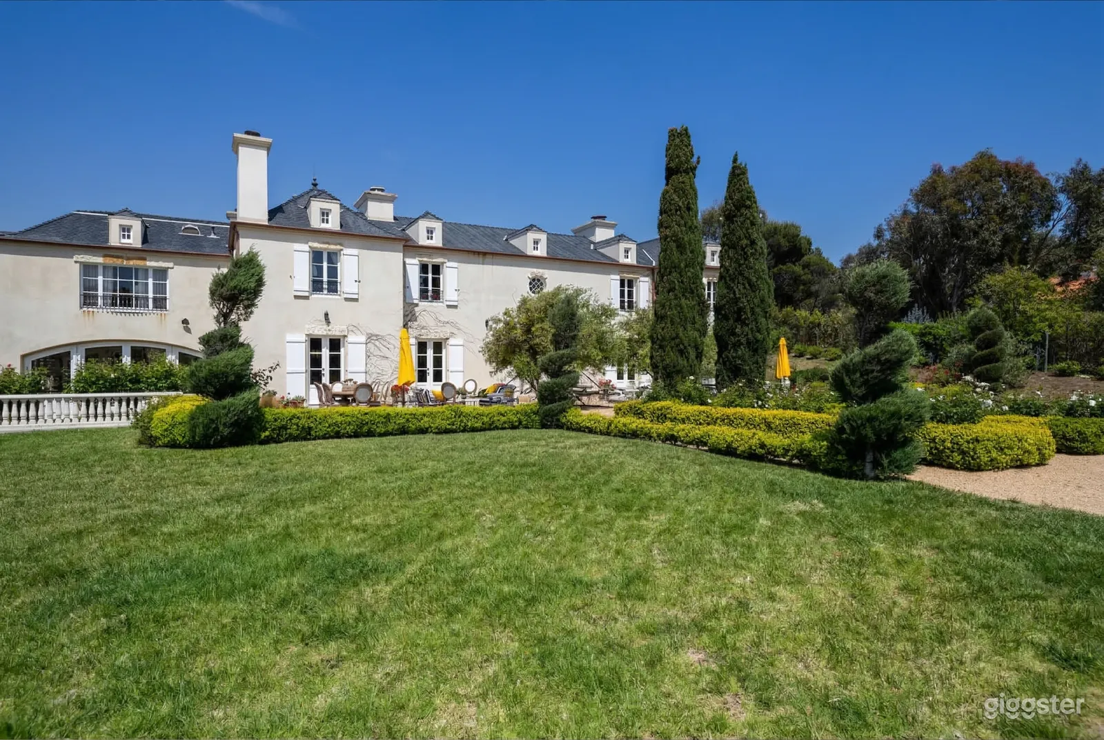 An expansive grey mansion overlooks a large garden with carved hedges and trees.