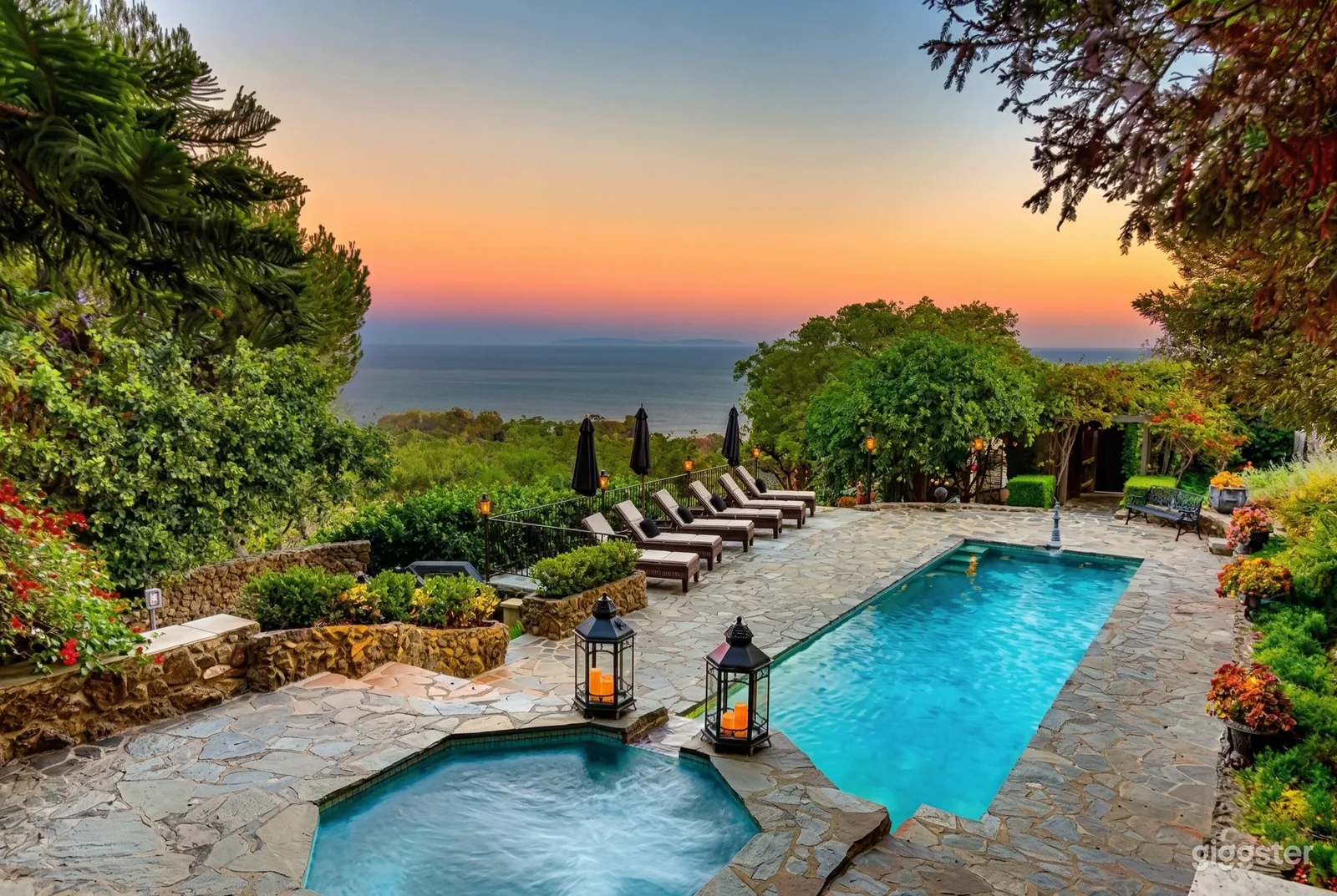 Lounge seating sits poolside in a lush gardened backyard overlooking water at sunset.