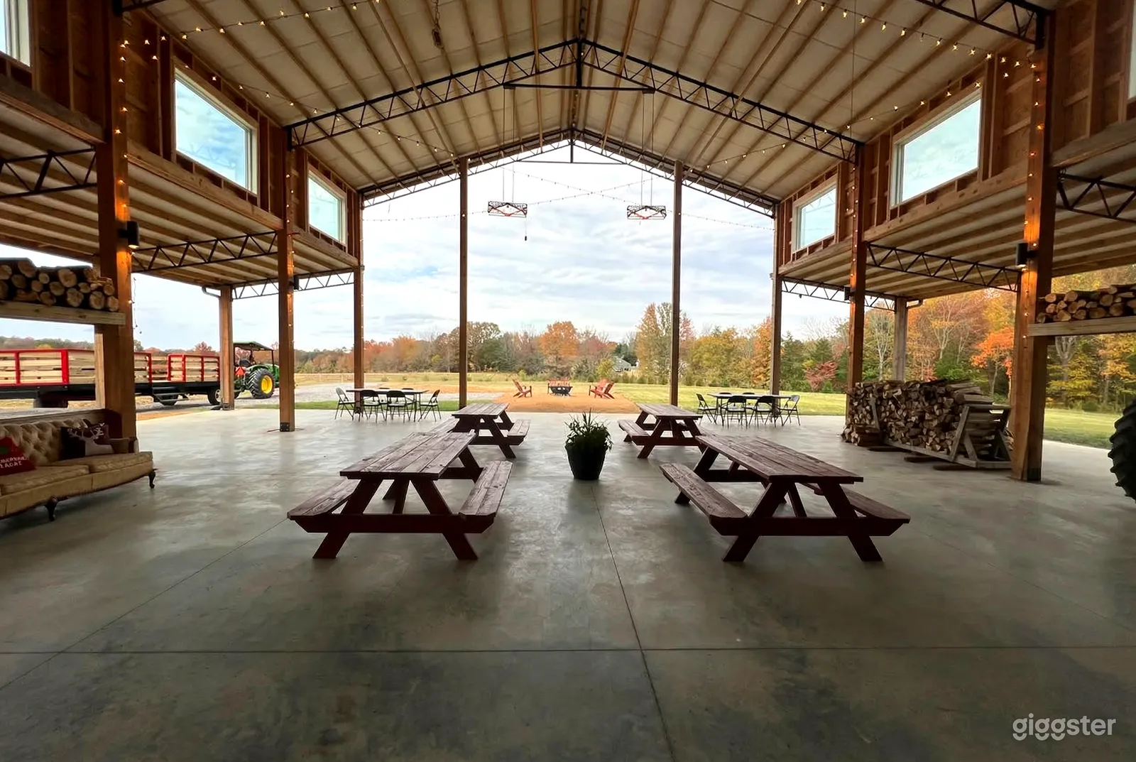 A large cement patio has four picnic tables and logs piled to the side under a metal roofed open building.