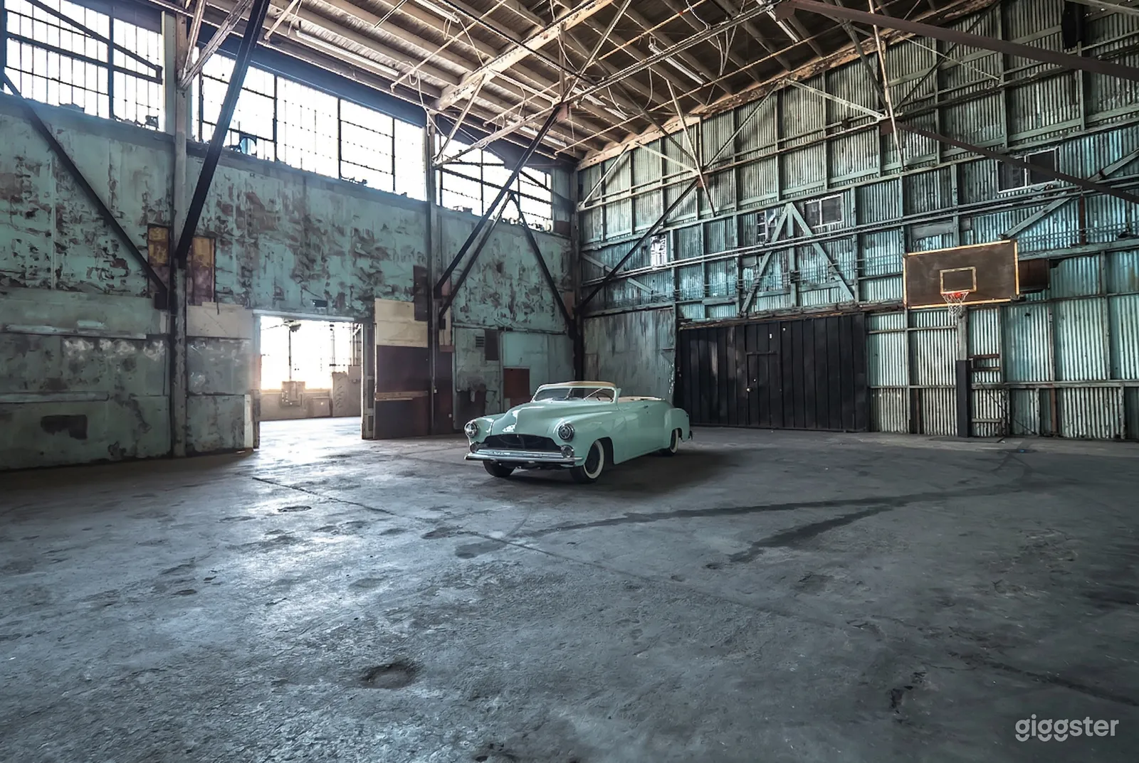 A turquoise vintage car sits in an expansive empty sound stage warehouse. 