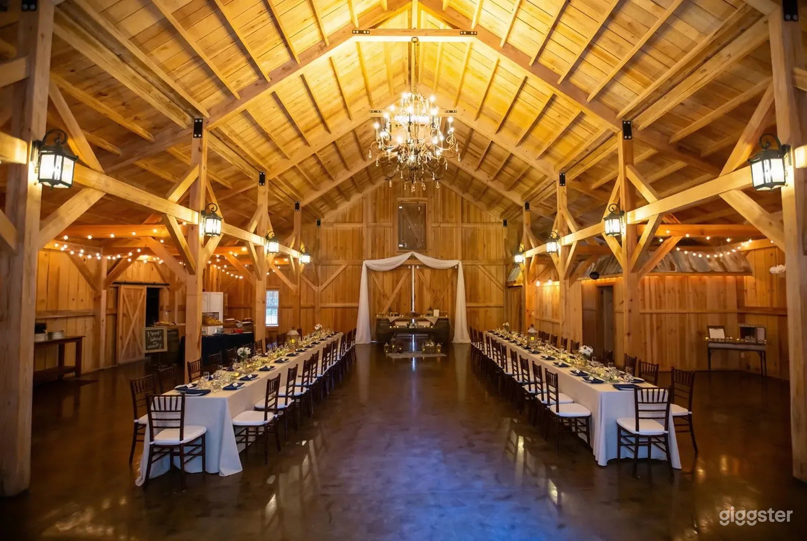 A rustic room with wood walls and ceiling has two long banquet tables set up and a focal point table with curtain draping at the front.