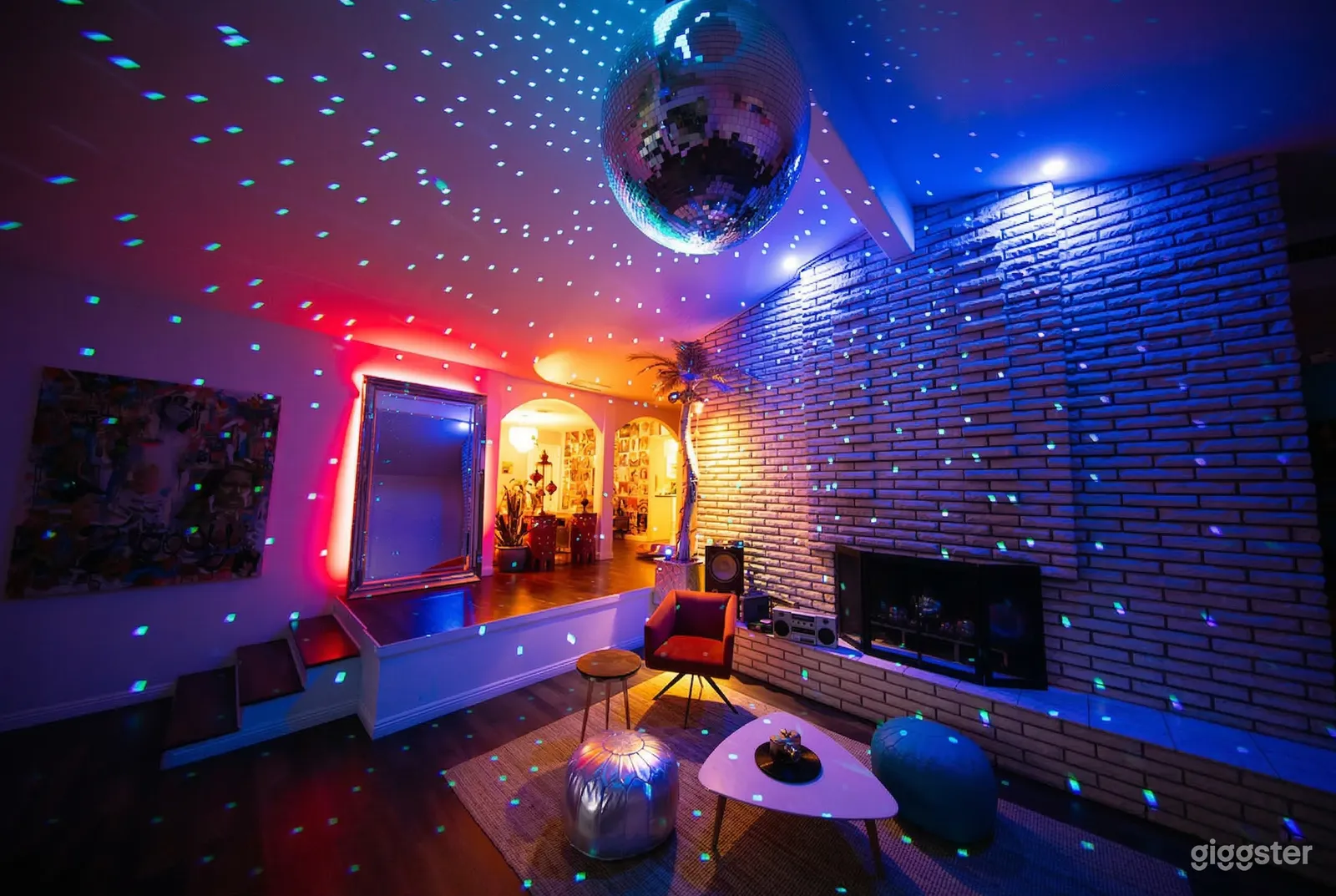 Varying chair styles surround a small table in front of a shiny brick wall in a room lit in violet and pink hues.