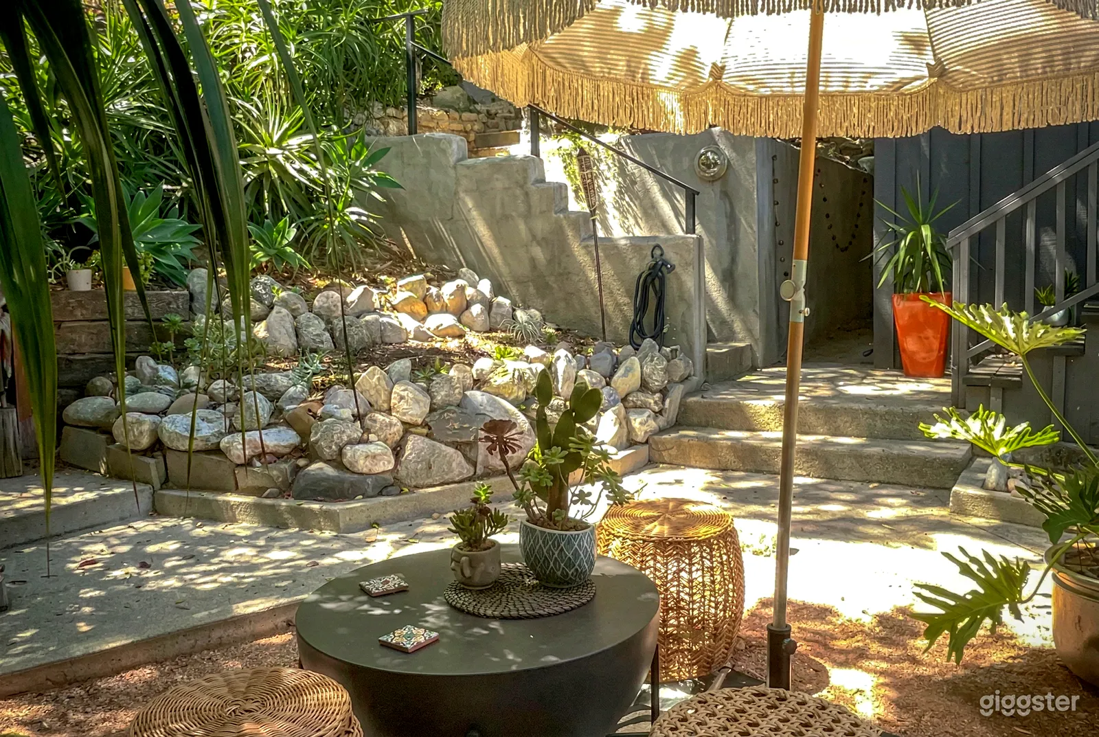 A table with wicker stools sits in front of cement stairs and a rock garden.
