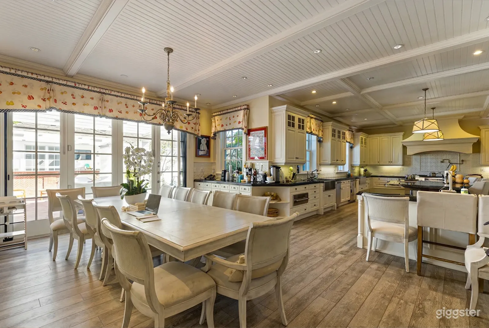 A large white dining table sits in front of a floor to ceiling window in a big homey kitchen.