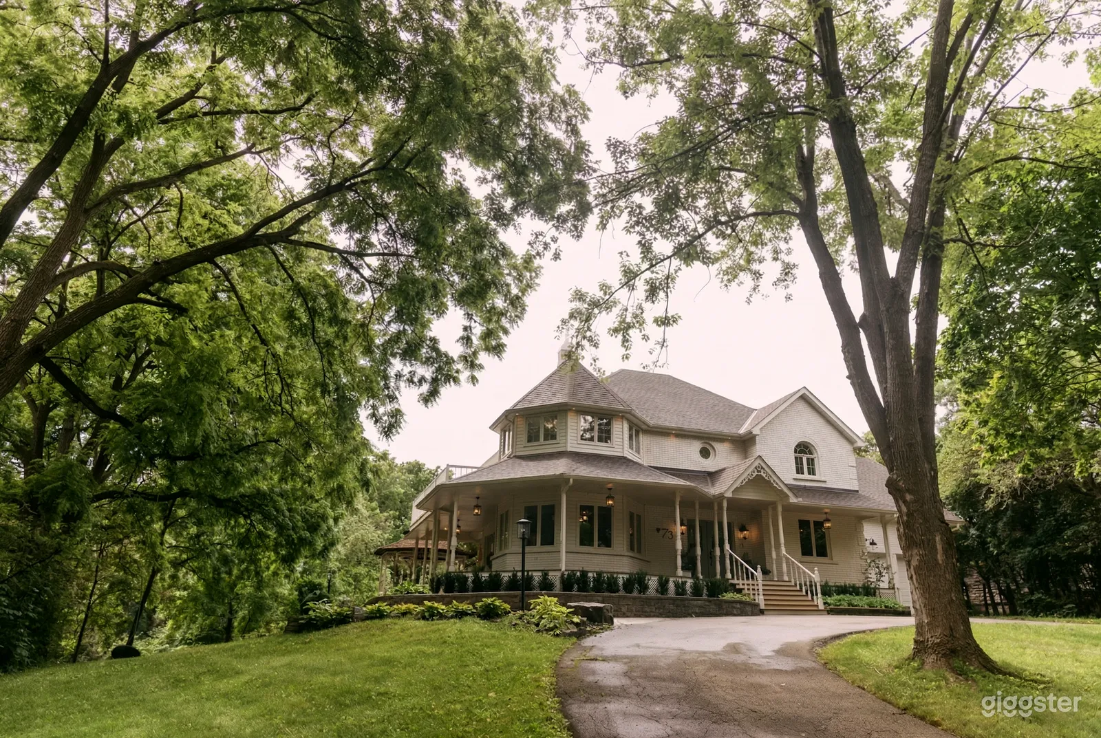 A large Victorian styled home with a wrap-around porch in a tall treed setting overlooking a circular drive.