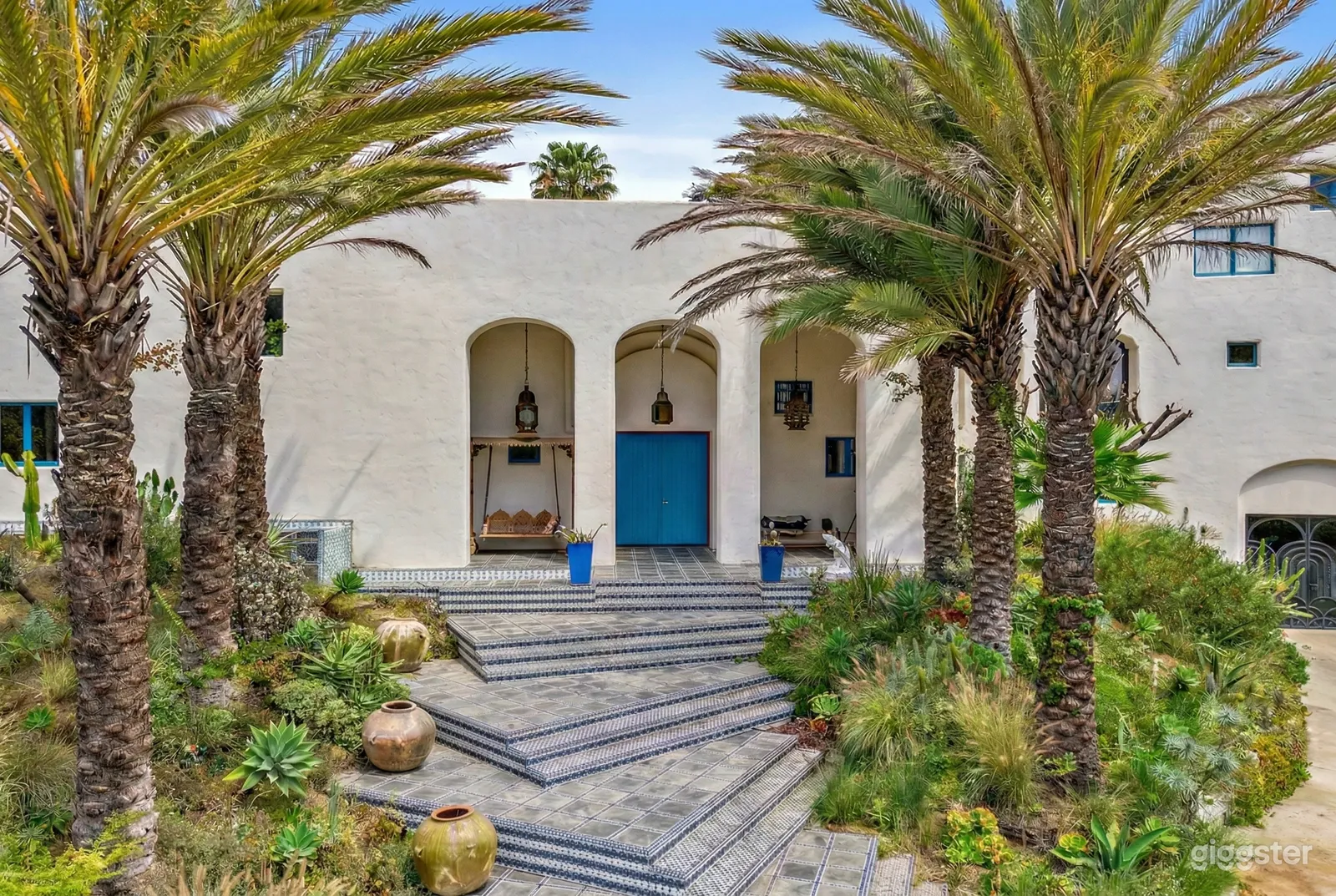 A white hacienda style building has a blue and white tile low stairway flanked by palm trees.