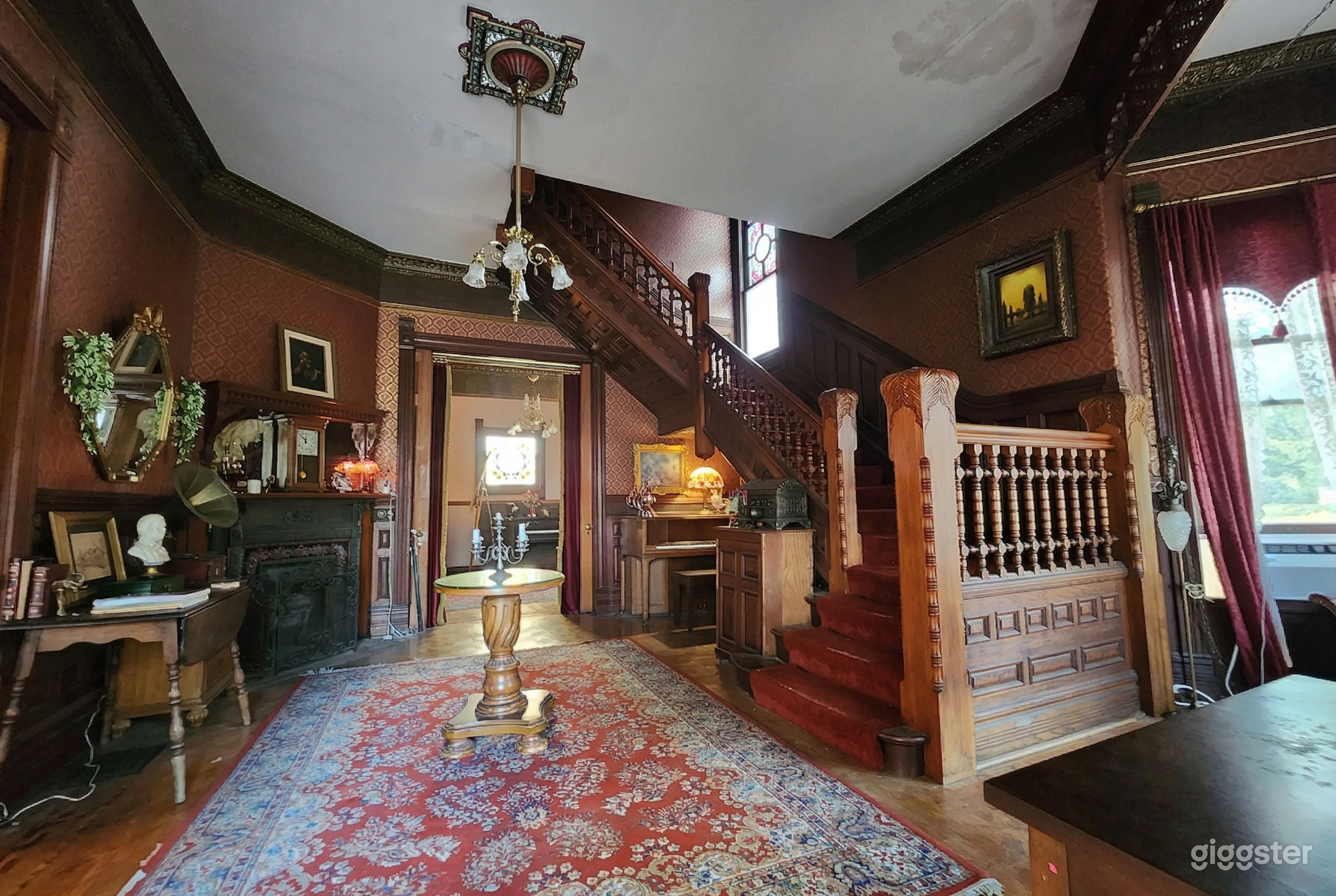 A big ornate staircase climbs to the upstairs from a main level historical home decorated with a vintage fireplace and tables.