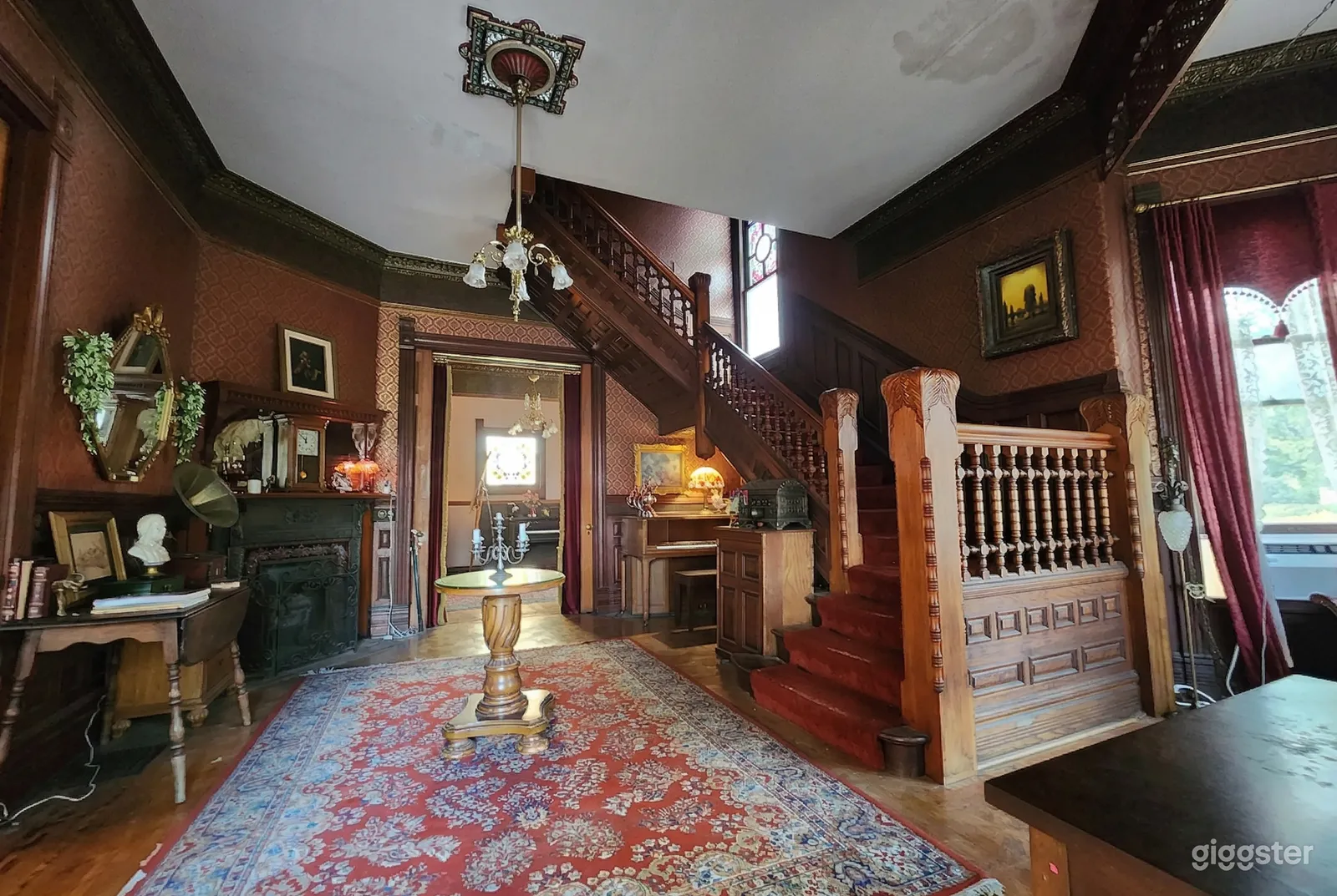A big ornate staircase climbs to the upstairs from a main level historical home decorated with a vintage fireplace and tables.