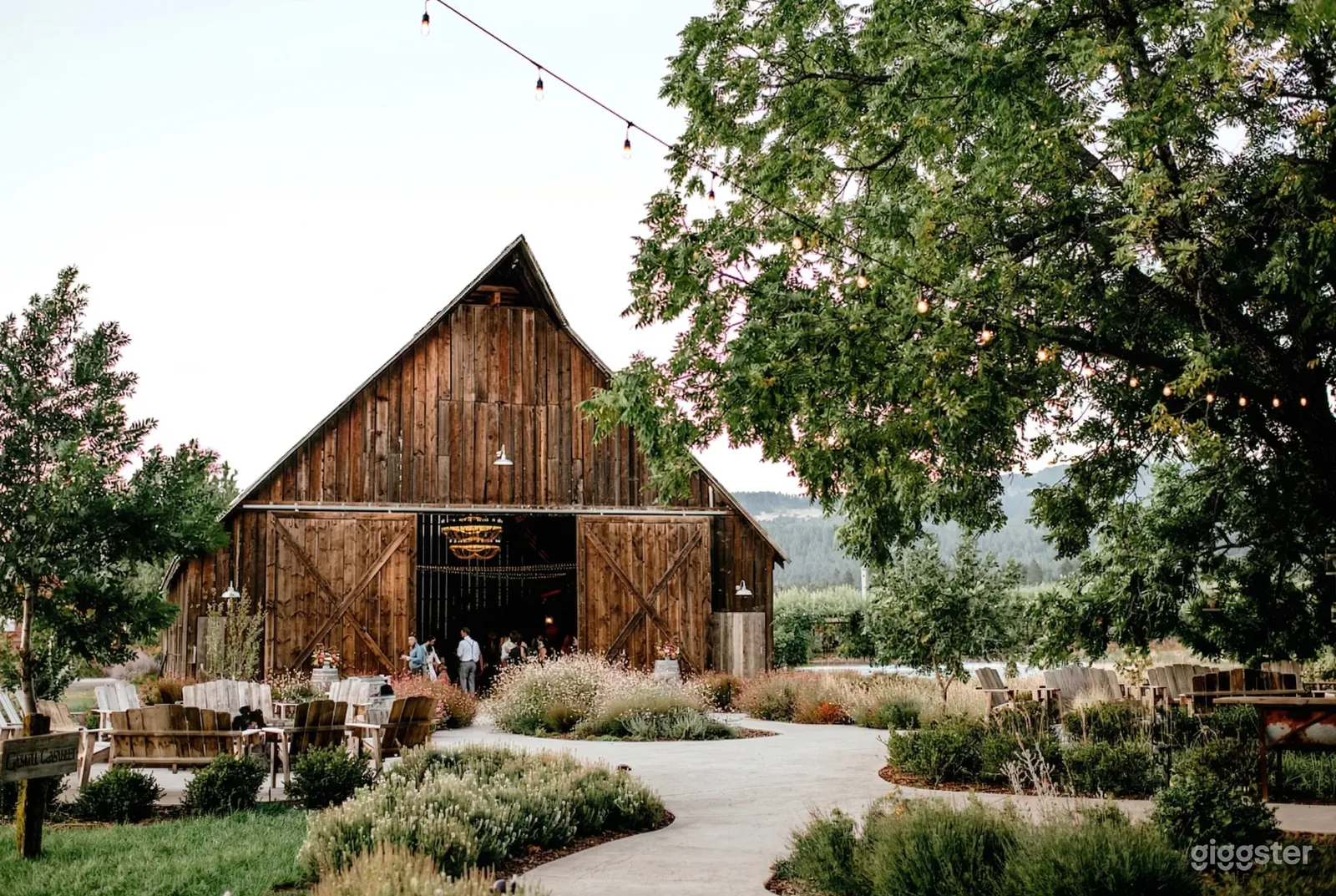 People entering a rustic barn nestled amongst trees and fields, with some seating in front and to the side.
