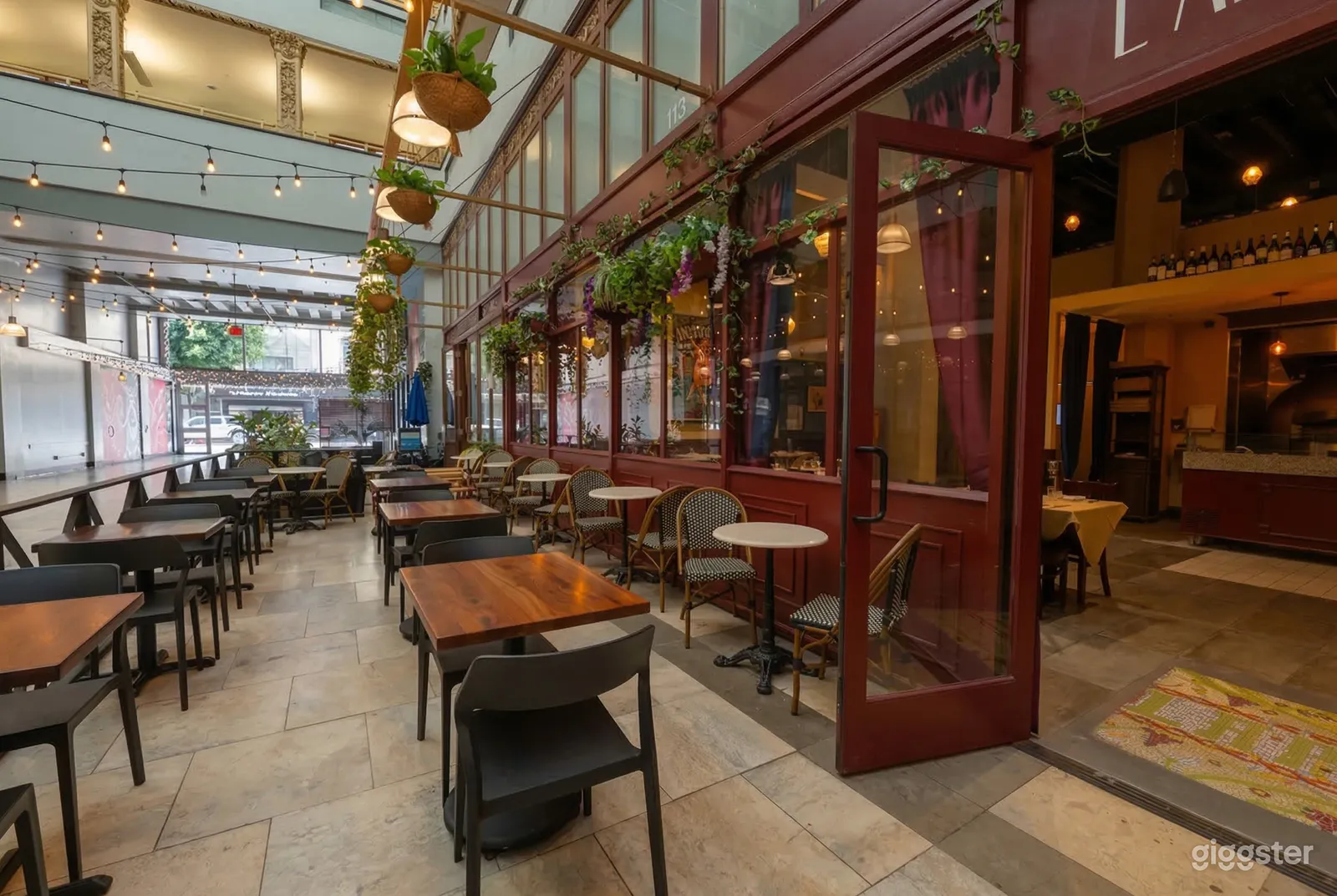 Rows of varying tables and chairs line a restaurant's indoor terrace area, with hanging plants and string lights overhead.
