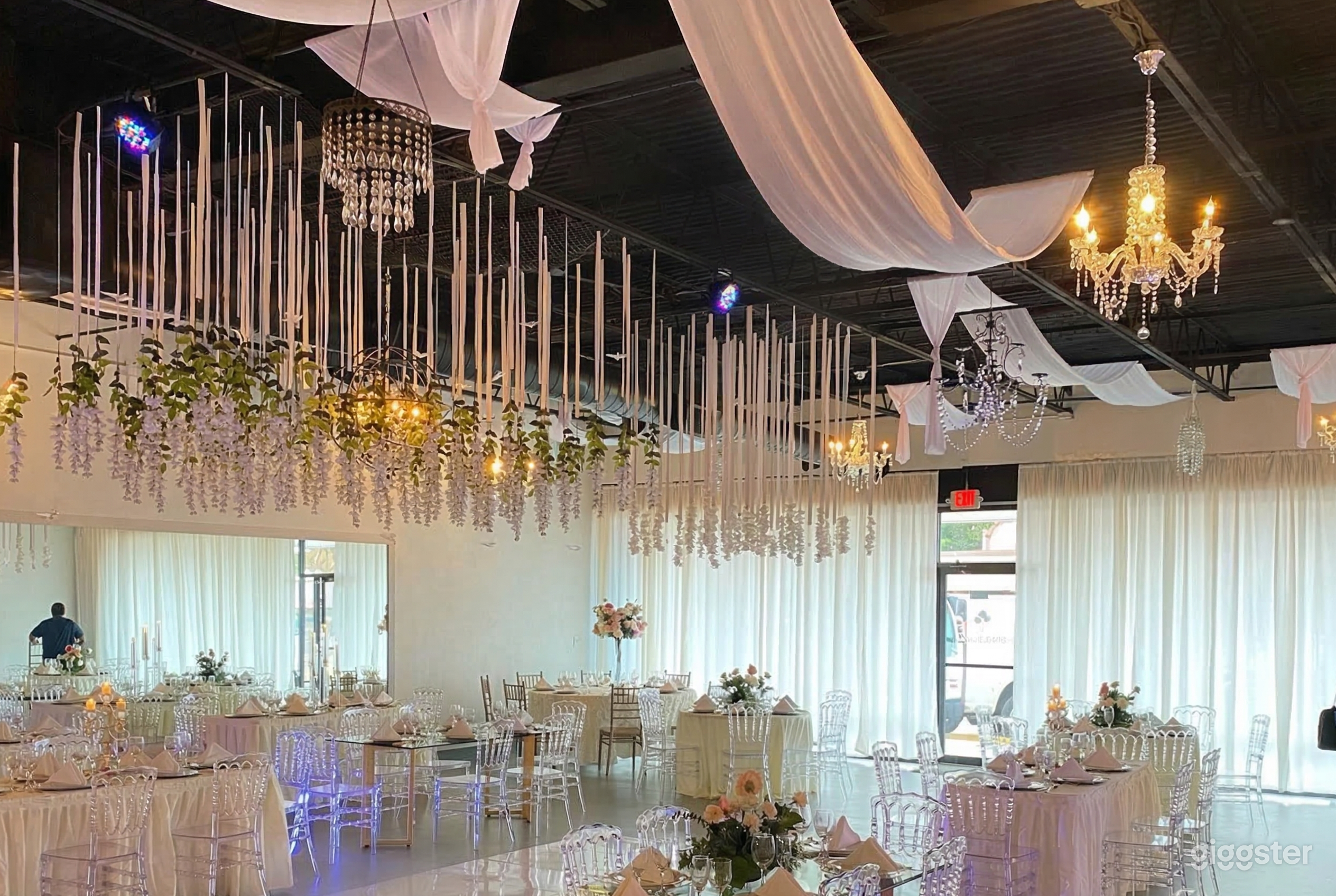 Tables with clear plastic chairs underneath a ceiling decorated with hanging florals and lighting.