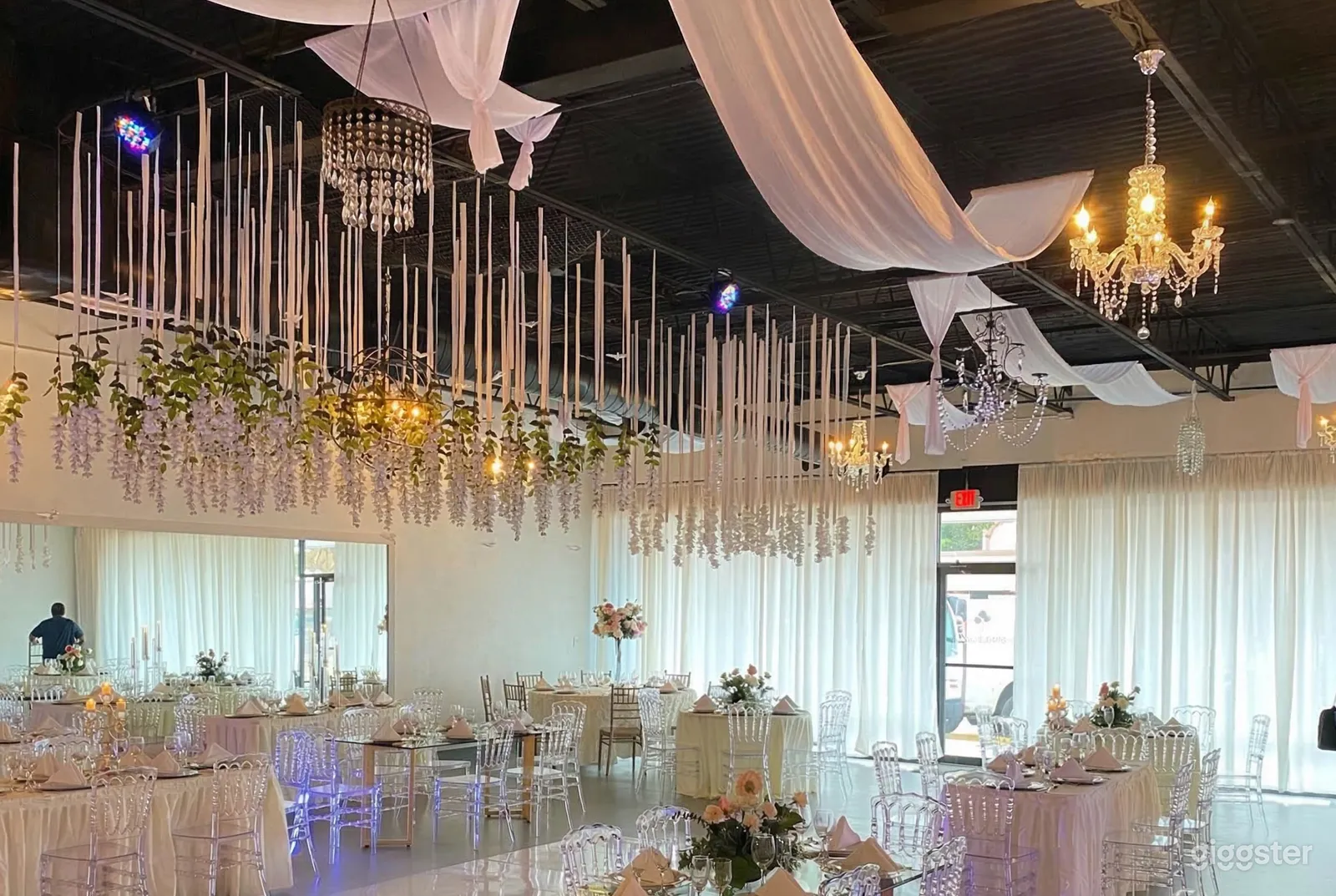 Tables with clear plastic chairs underneath a ceiling decorated with hanging florals and lighting.