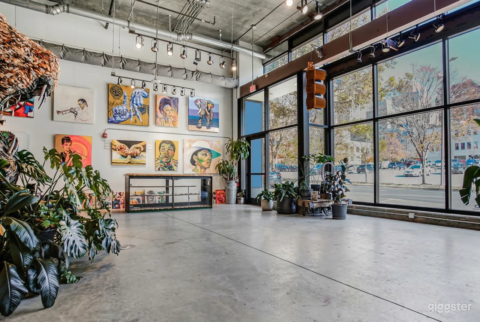 A large warehouse type event room is decorated with large painting and plants, and has floor to ceiling windows.
