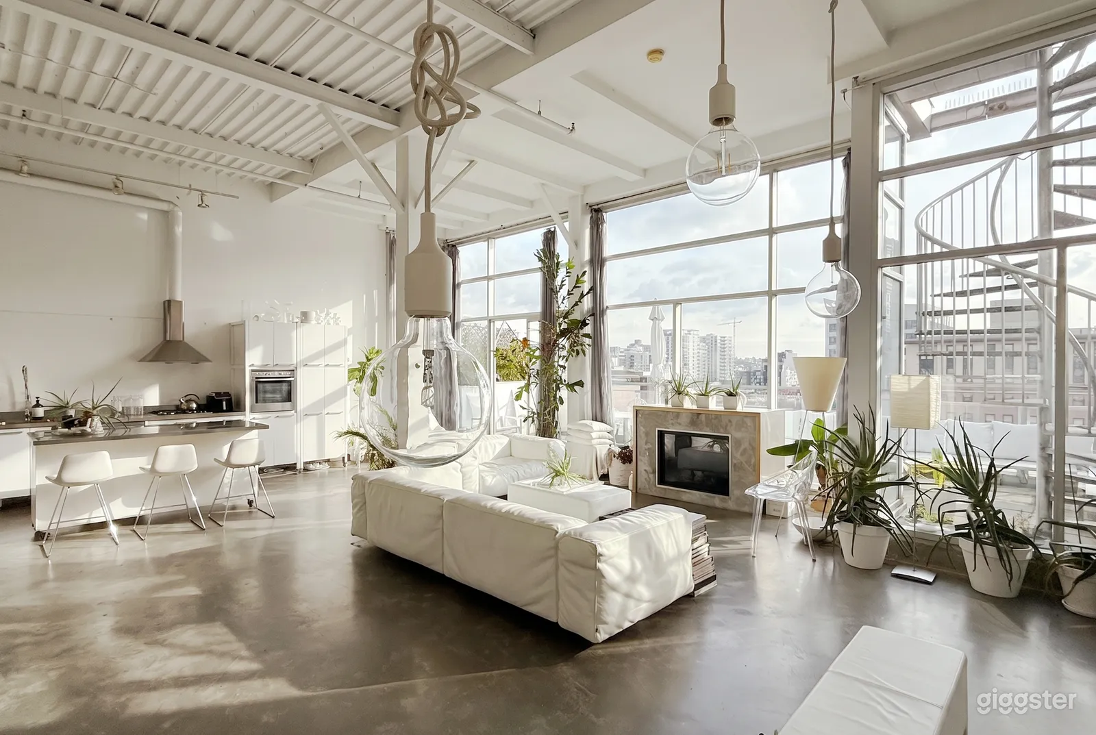 A room with a wall of windows, cement floor, white furniture, a kitchen area and large industrial bulb lighting hanging down.
