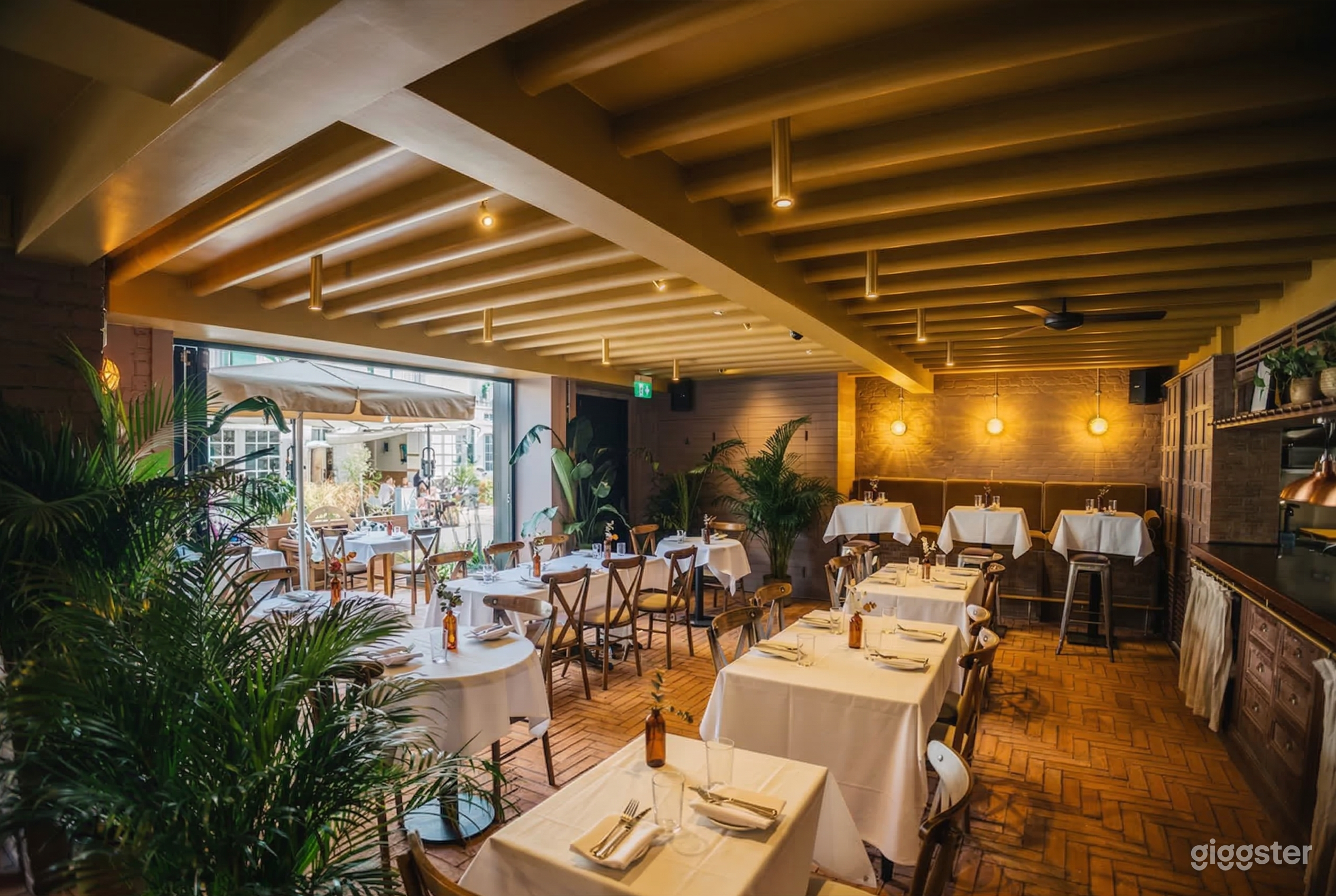 Multiple shaped tables covered in white in a well-lit restaurant and terrace decorated with plants.