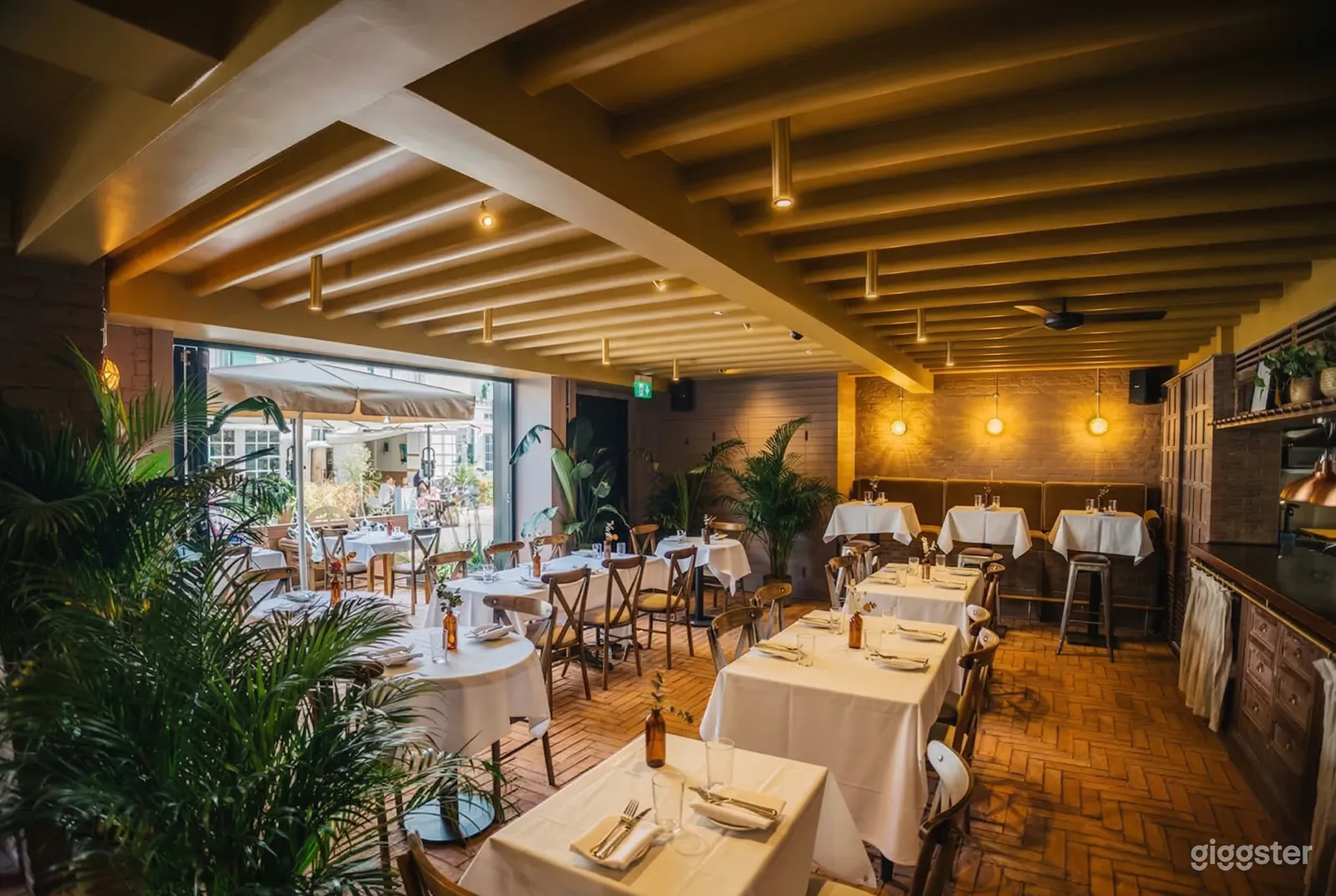 Multiple shaped tables covered in white in a well-lit restaurant and terrace decorated with plants.