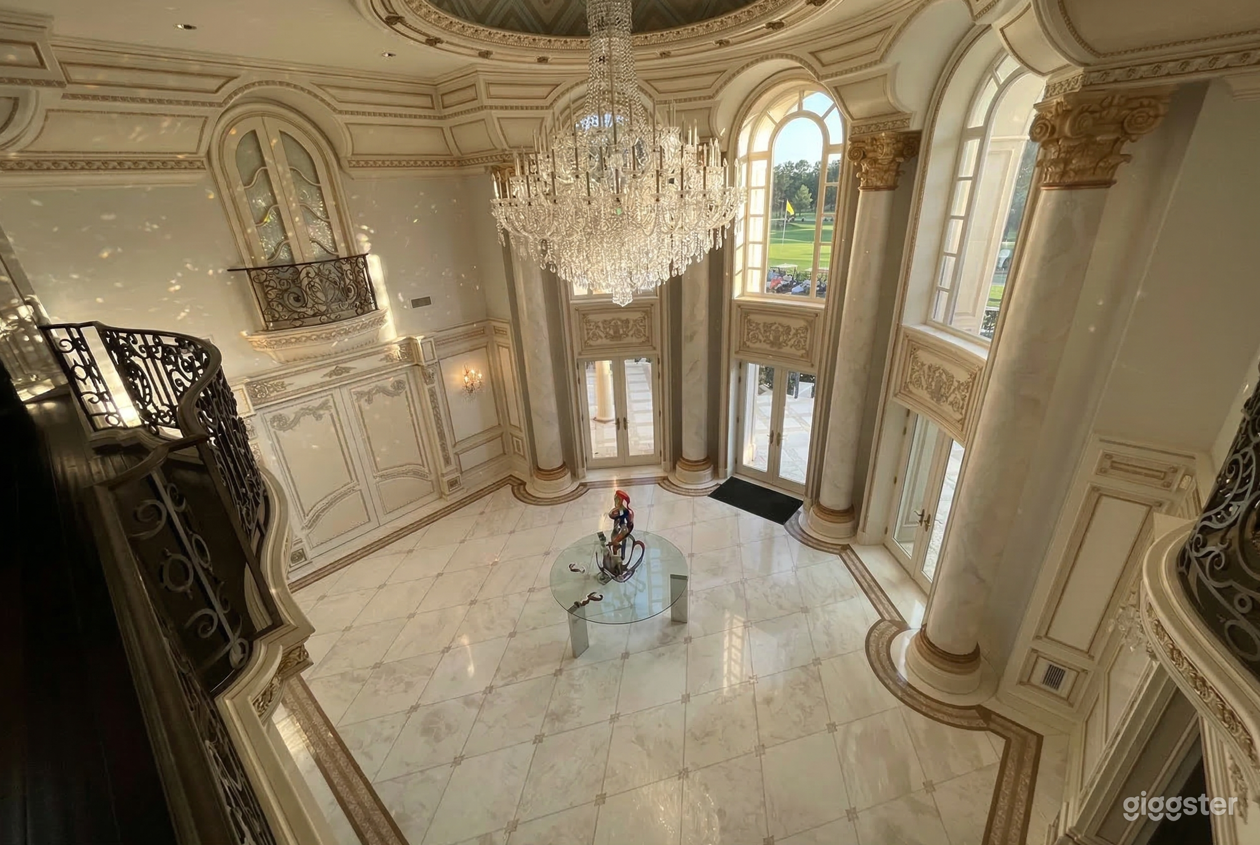 A chandelier hangs over a glass table centered in an ornate room with stone columns, high windows, and balcony with wrought iron.