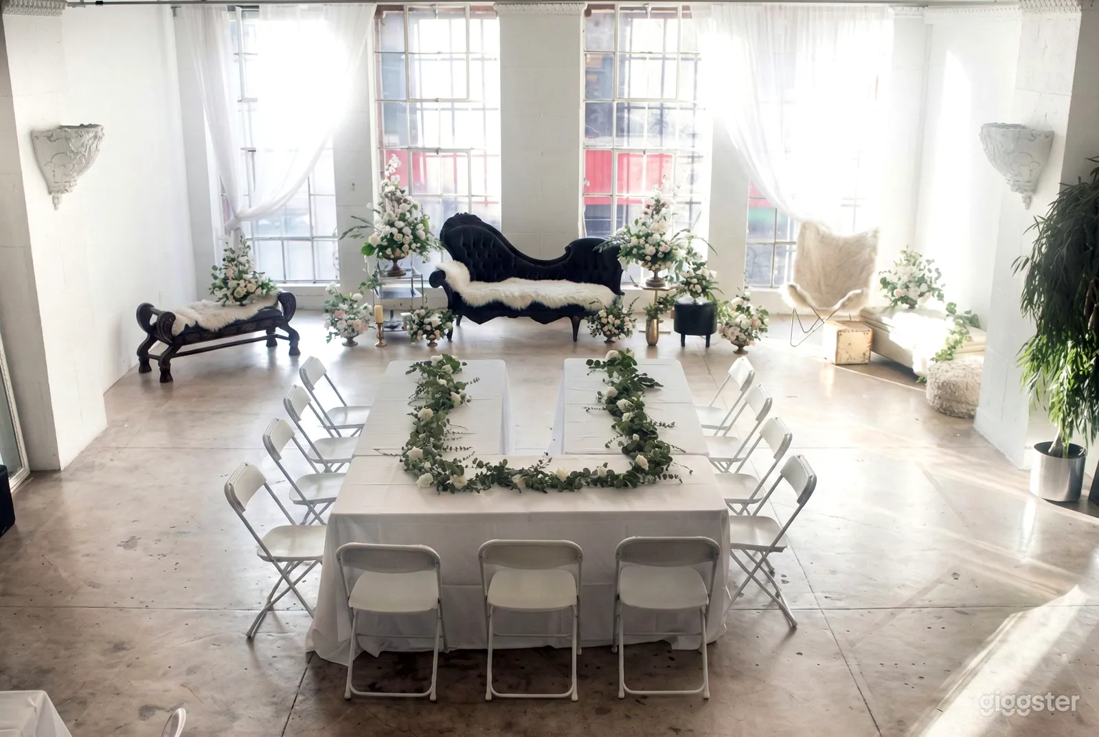 A white u-shaped table decorated with greenery in front of a settee flanked by florals.
