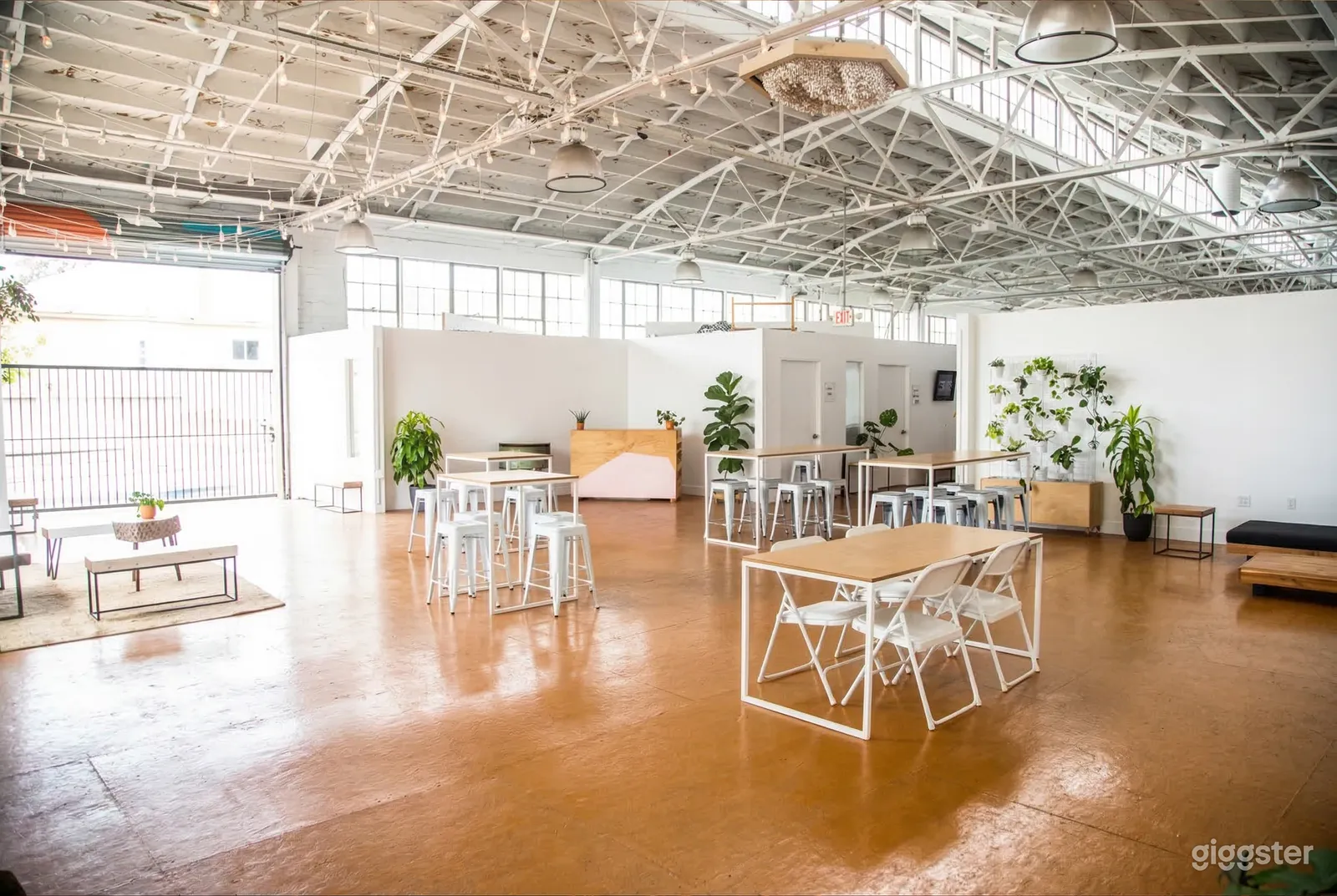 A white open-space industrial style room contains wood and white tables and white chairs and has a ceiling with white iron detail.