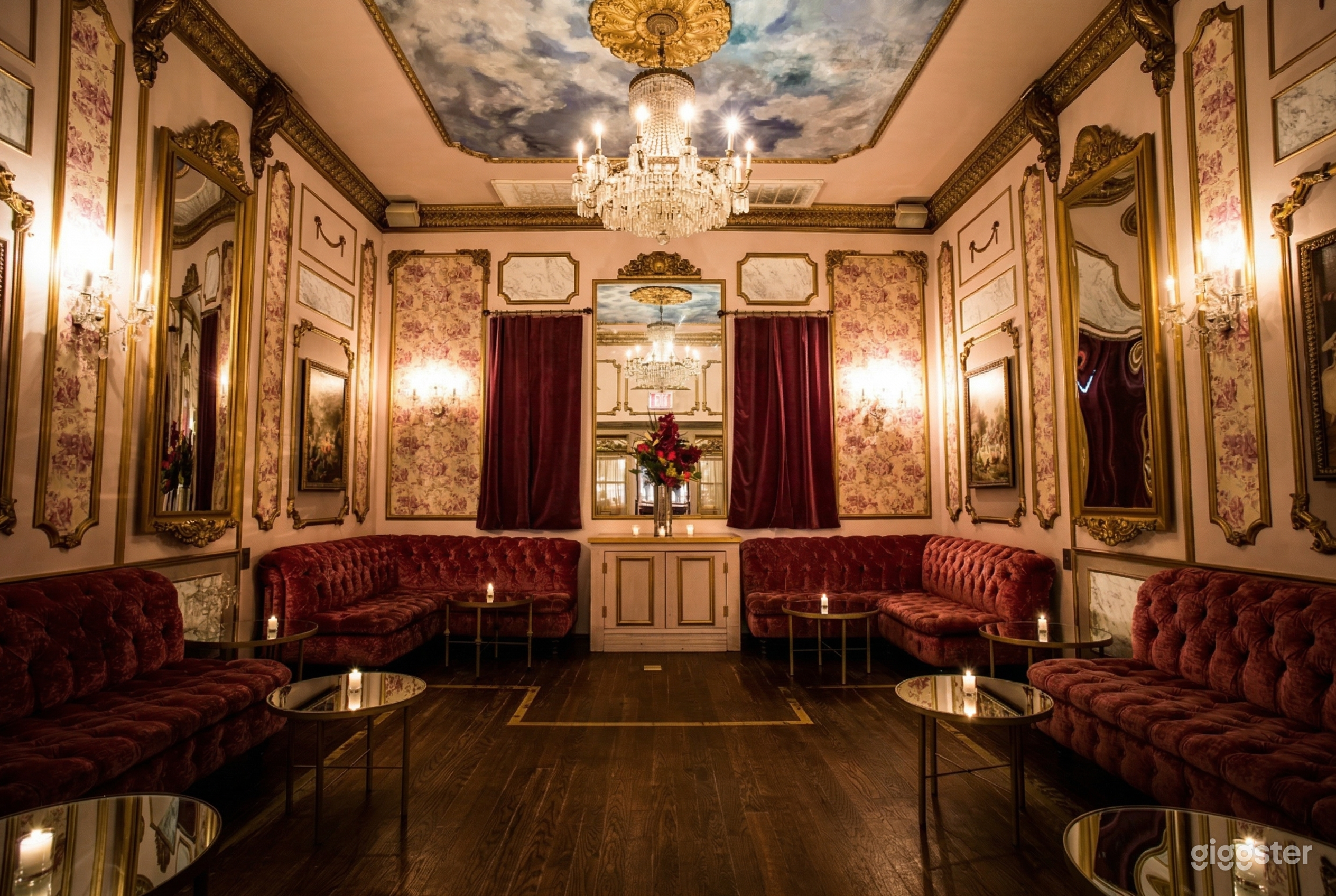 A Victorian style room with floral wall paper, red velvet furniture and curtains, and a central ceiling chandelier.