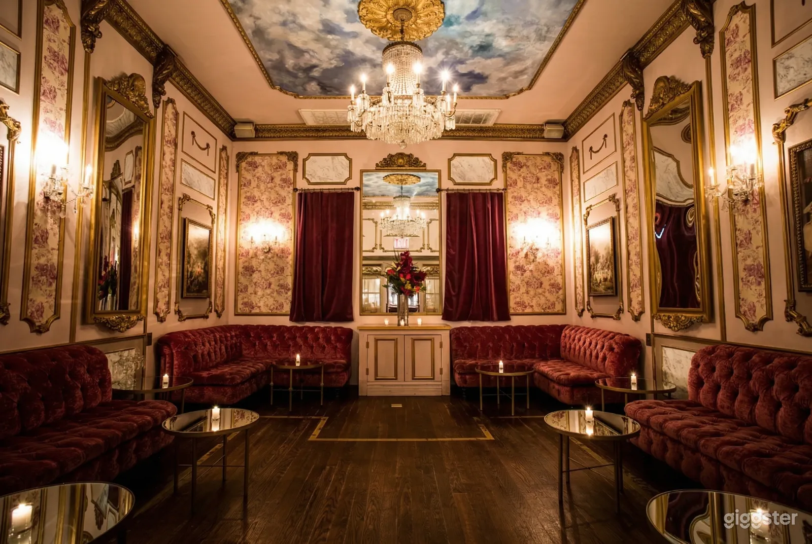 A Victorian style room with floral wall paper, red velvet furniture and curtains, and a central ceiling chandelier.