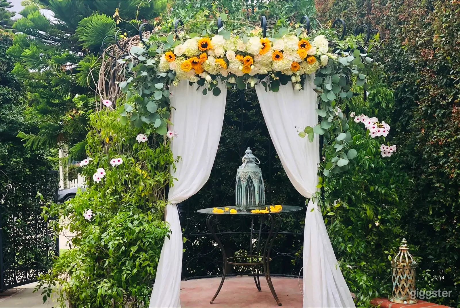 A small black table is centered under a floral and greenery arch and flanked by white sheer curtains.